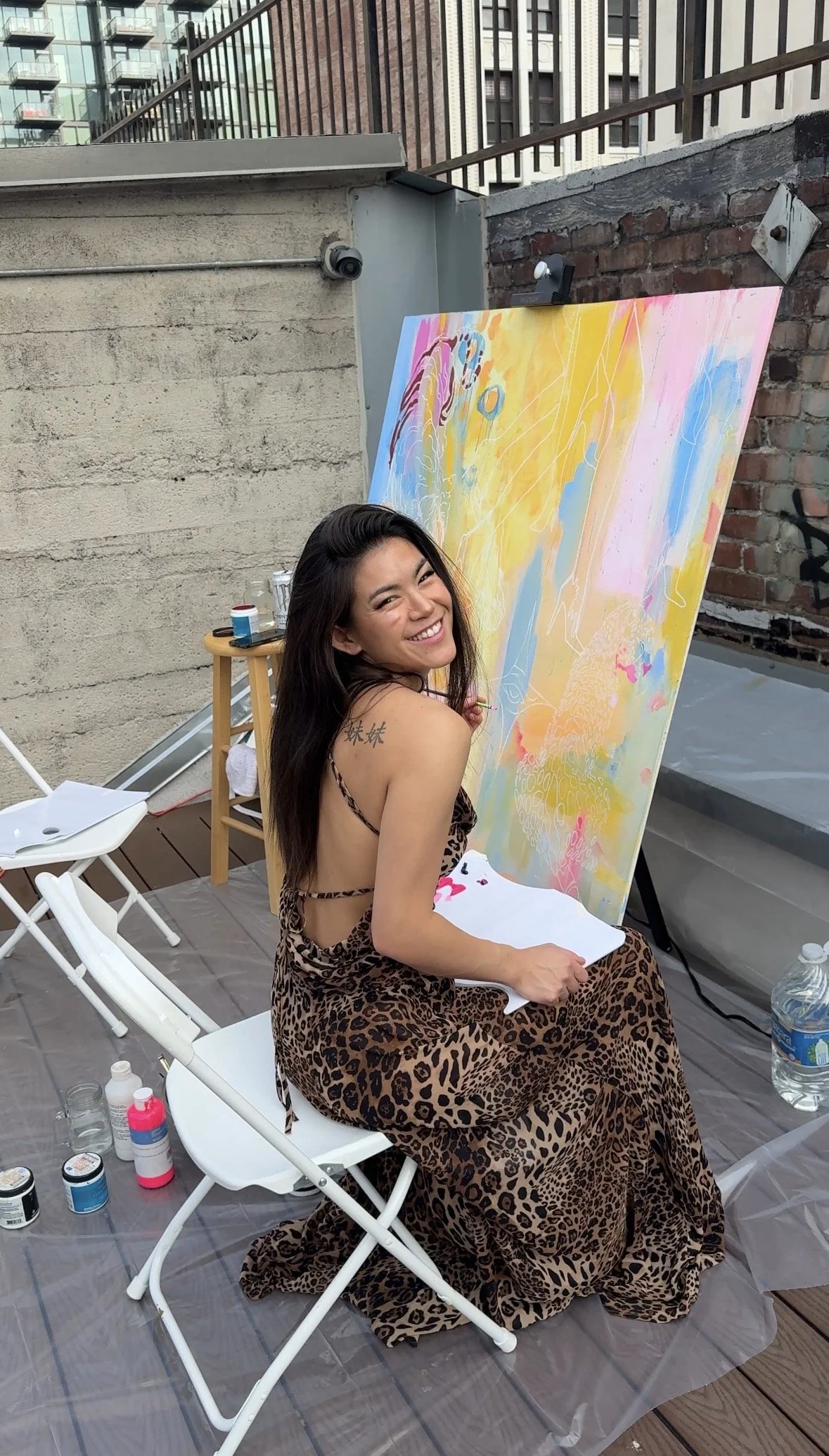 A woman wearing a leopard print dress is sitting on a deck chair, smiling at the camera, with a large colorful abstract painting behind her. Art supplies and bottles are around her, and there is a plastic sheet under her.
