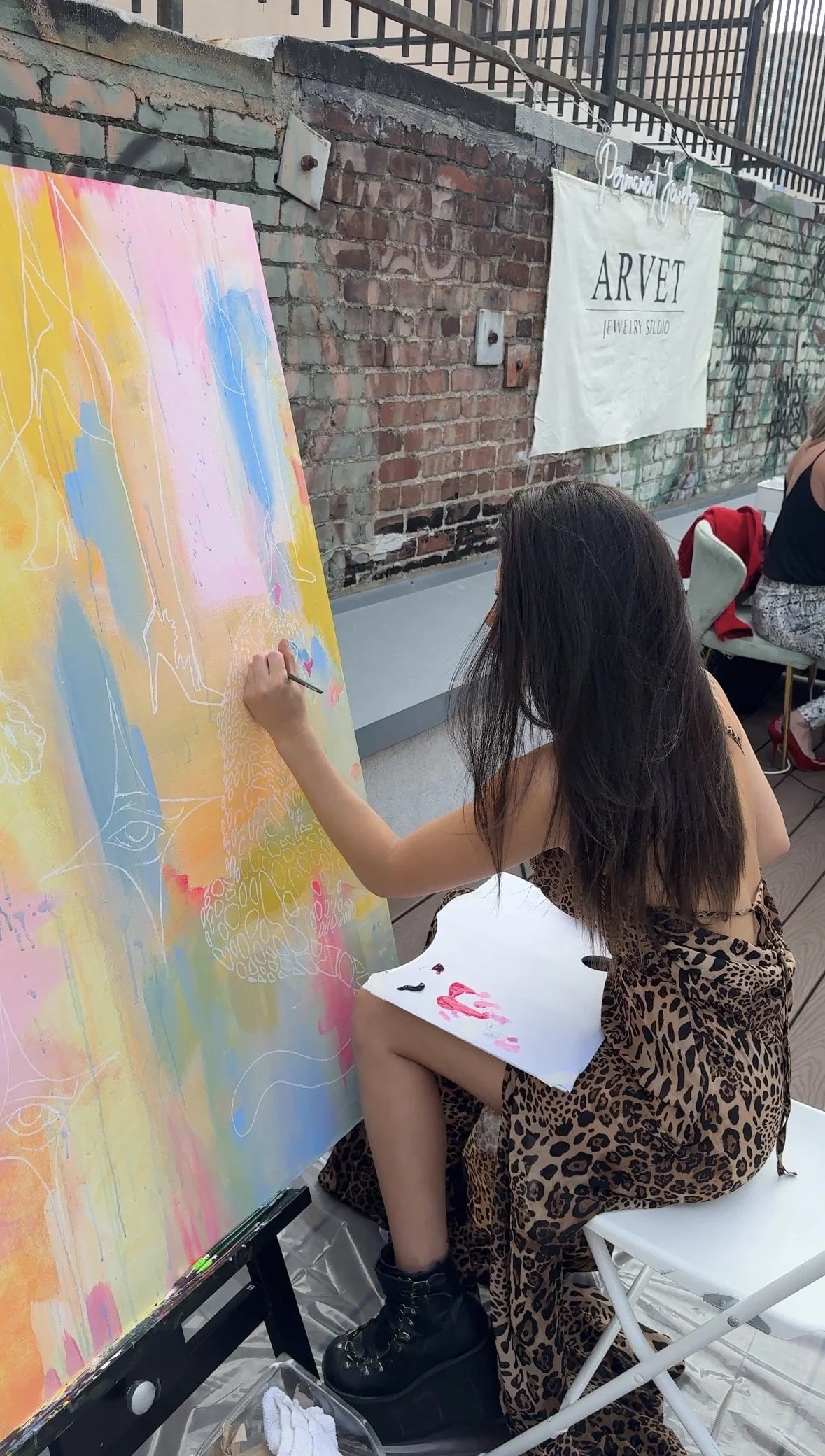A young woman in a leopard print dress and black boots is painting on a large canvas with pastel colors, abstract lines, and patterns. She is sitting on a white folding chair, working outdoors against a brick wall with graffiti and a white banner tha