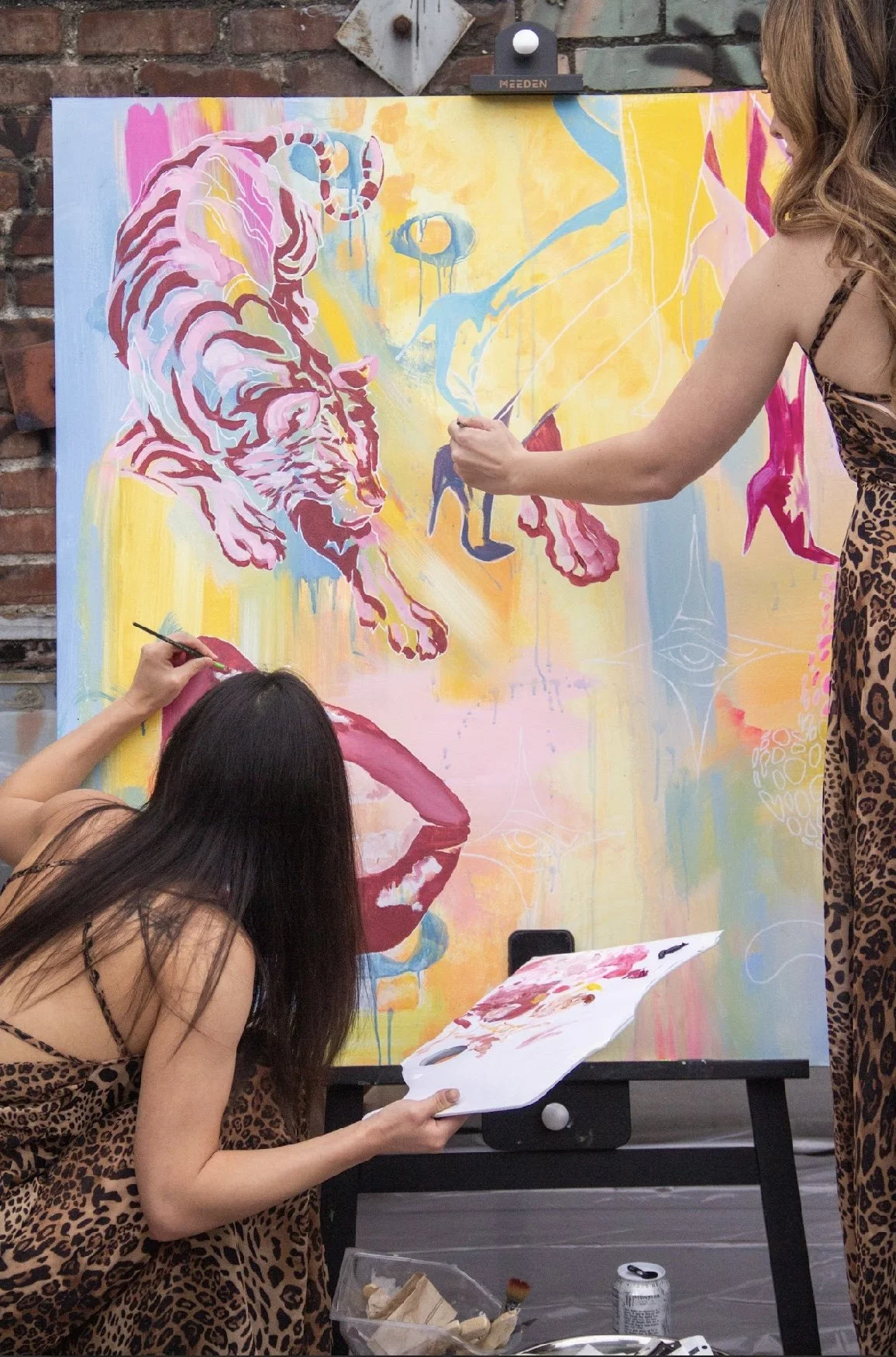 Two women painting a colorful, abstract mural on a large canvas against a brick wall. One woman with long dark hair is kneeling, holding a palette and brush, while the other with long brown hair wearing a leopard print dress is standing, using a brus