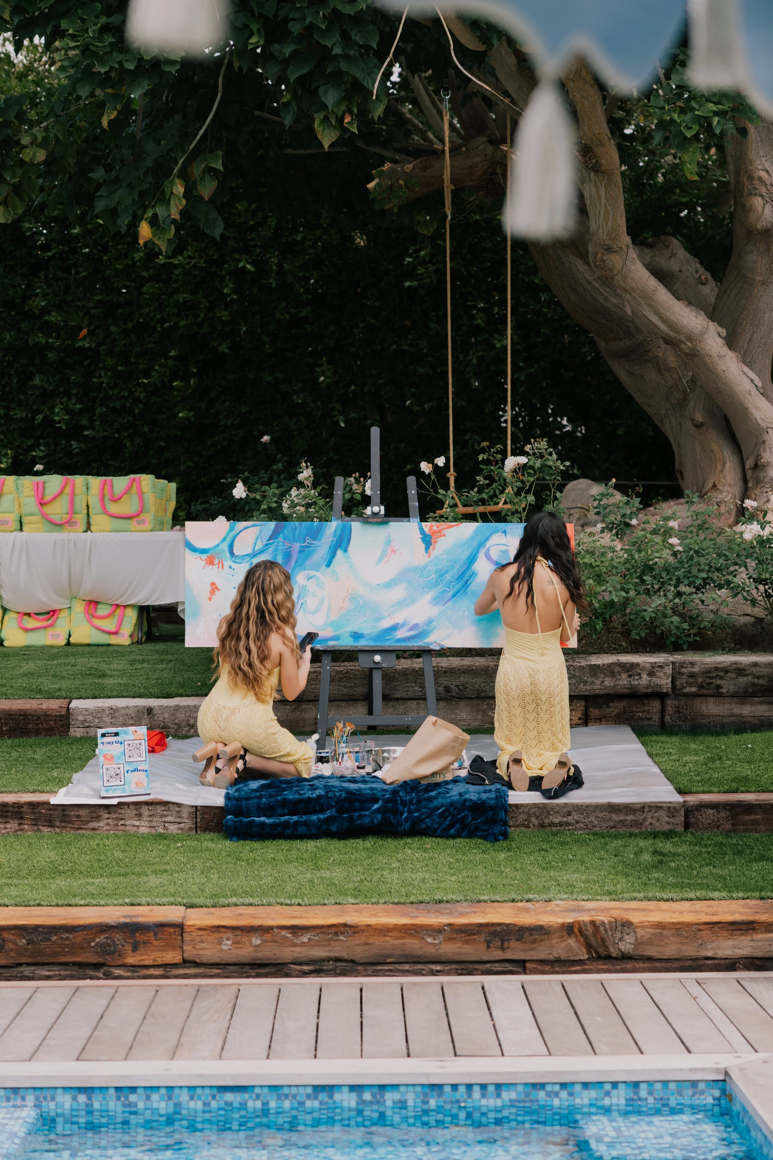 Two women dressed in yellow dresses sitting on a white mat in a backyard, painting a large abstract canvas on an easel, with a swimming pool in the foreground and a large tree in the background.