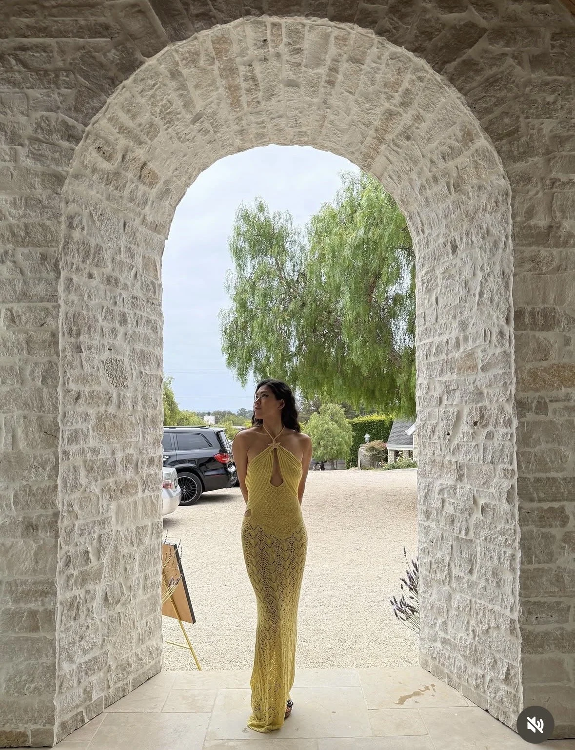 A woman in a yellow dress standing under a stone archway with trees and parked cars visible in the background.