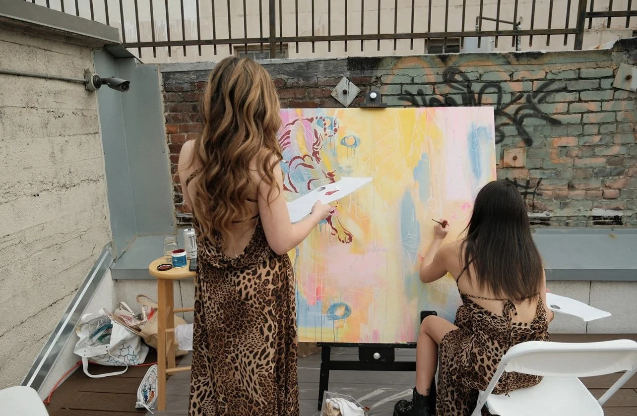 Two women with long hair wearing leopard print dresses painting on a large canvas outdoors.