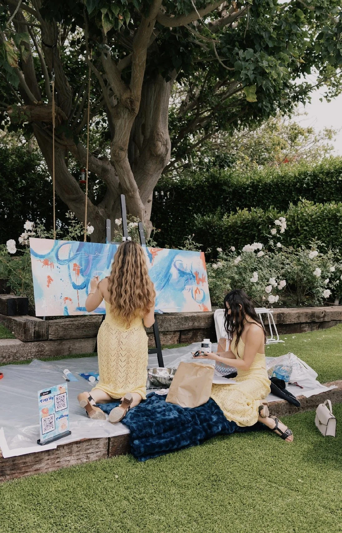 Two women in yellow dresses creating abstract art on a large canvas in a garden with trees and white flowers, with art supplies on a blanket on the grass.