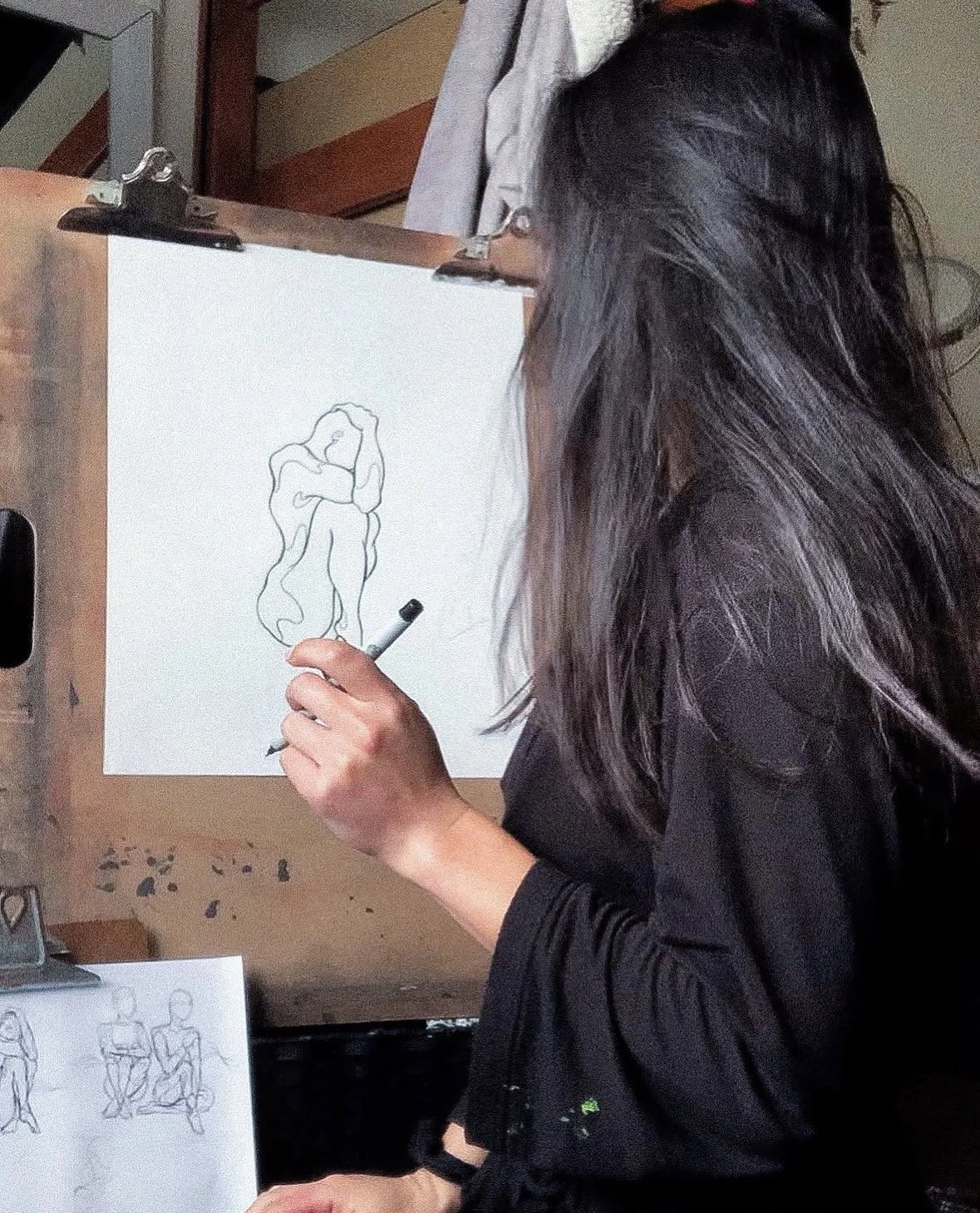An artist drawing a line art of a romantic couple embracing on a white sheet of paper, with additional sketches of seated figures visible nearby.