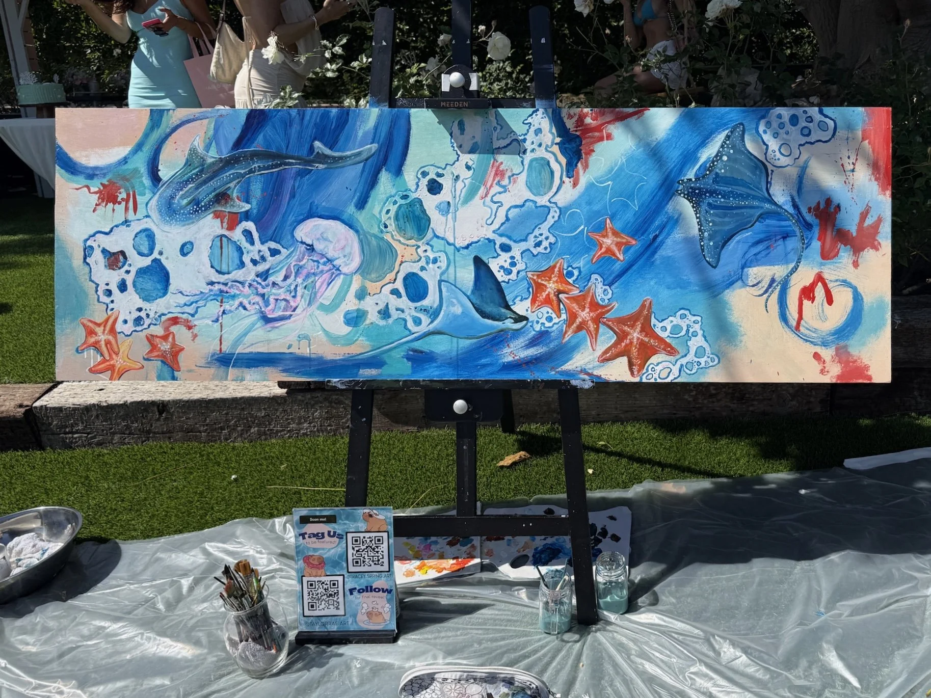 An outdoor painting of marine life, including rays, a jellyfish, and starfish, with vibrant blue, red, and white colors. The painting is on an easel, surrounded by art supplies and informational cards.