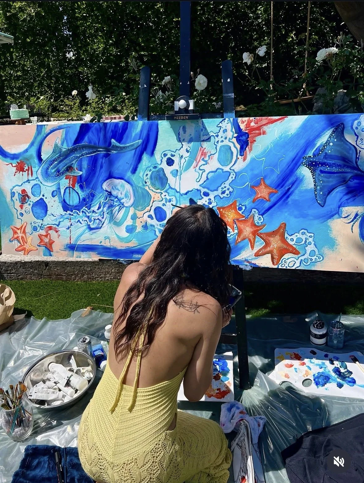 A woman with long dark hair in a yellow dress painting a large, colorful ocean-themed mural outdoors. The mural features sea creatures like sharks, starfish, jellyfish, and octopuses in vibrant blue, orange, and red tones. The woman is sitting on a p