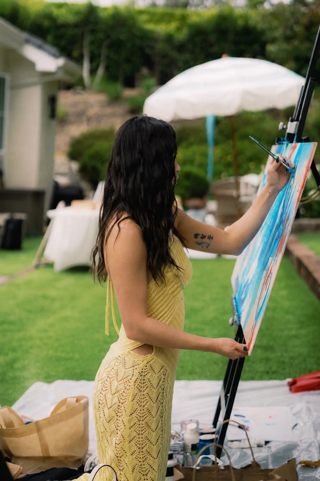 A woman in a yellow dress painting on a canvas outdoors with a brush, standing near a picnic table and a white umbrella.
