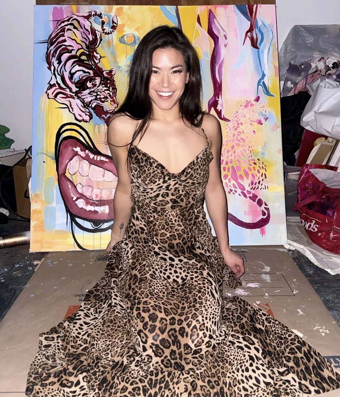A woman wearing a leopard print dress smiling at the camera, sitting in front of a colorful painting of a tiger's face and a pair of legs with painted toes.