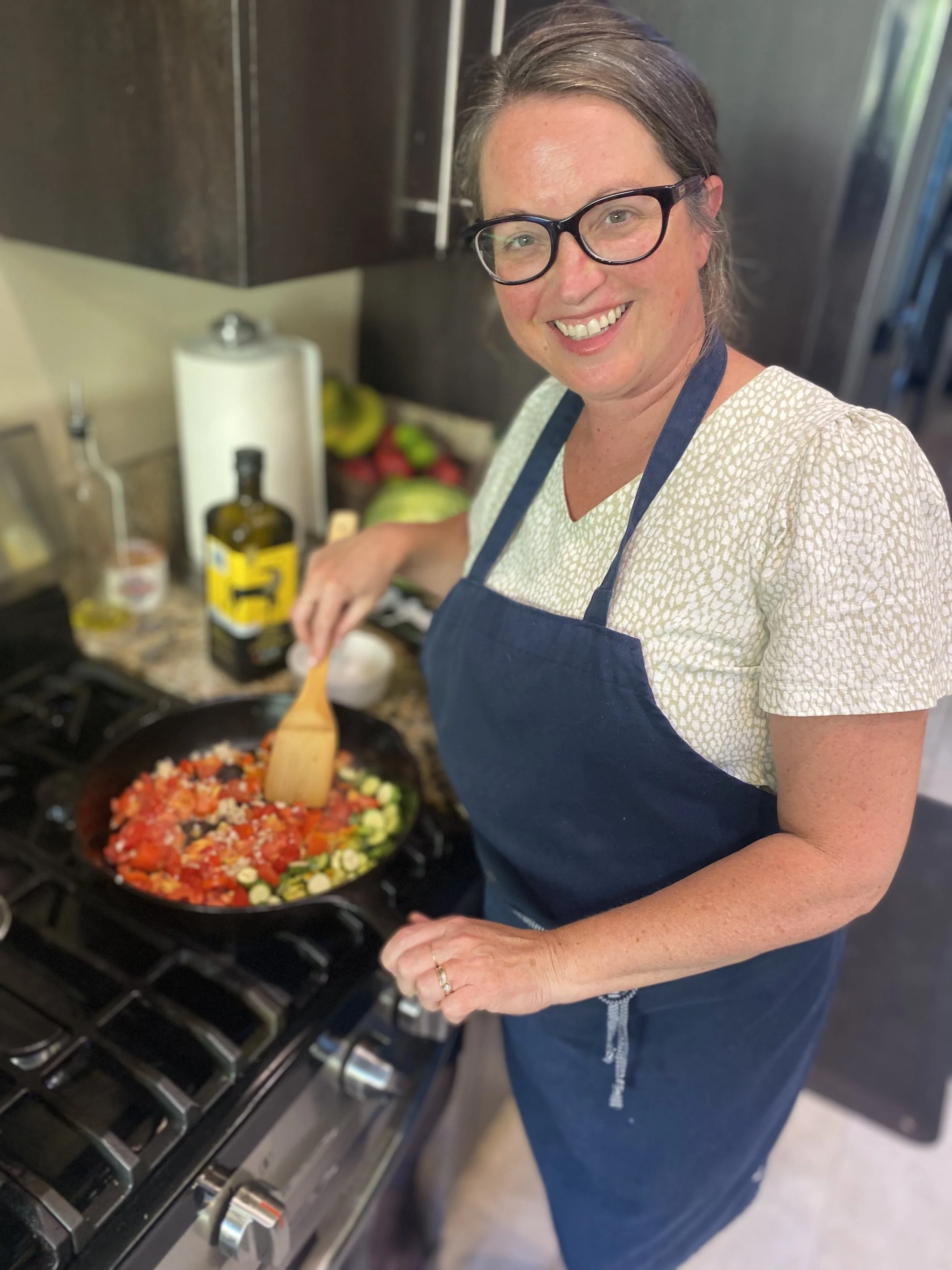 A woman wearing glasses, a light-colored patterned shirt, and a dark apron is cooking vegetables in a frying pan on a stove. She is smiling and looking at the camera.