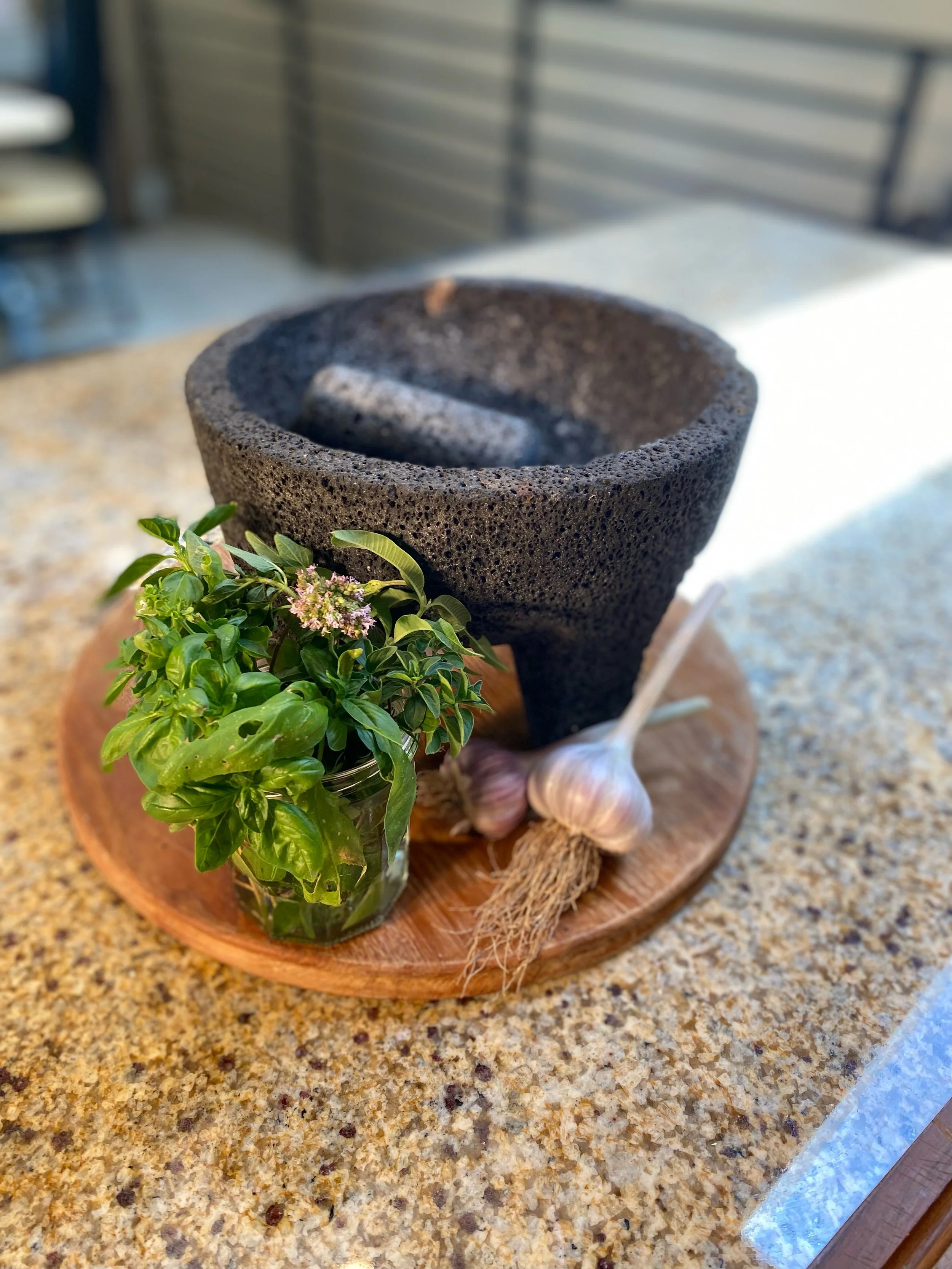 A potted herb plant with green leaves and small pink flowers, a bunch of garlic bulbs, a garlic piece, and a black stone mortar on a round wooden tray on a speckled countertop.
