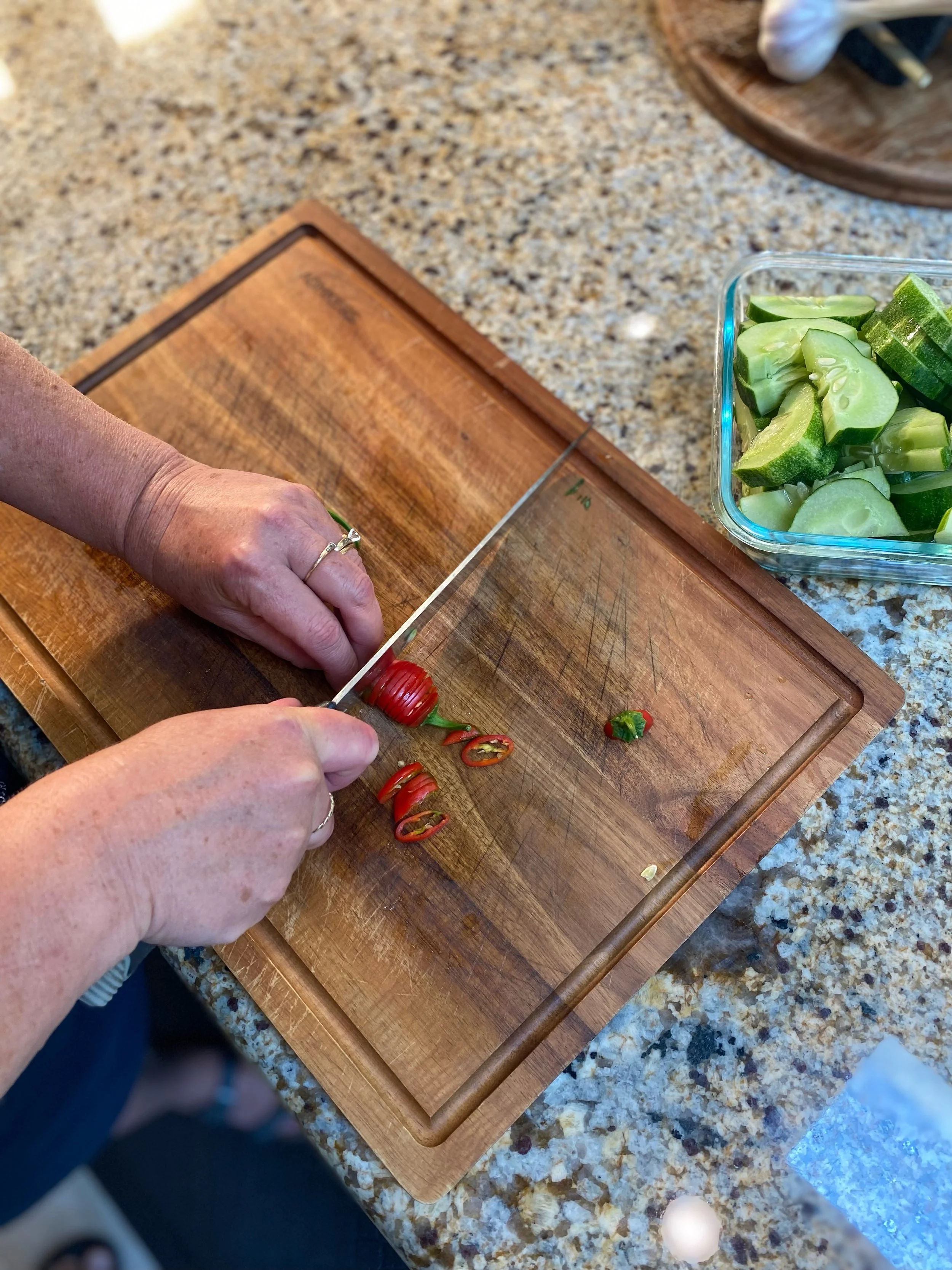 A person is slicing red chili peppers on a wooden cutting board with a knife, beside a container of sliced cucumbers on a kitchen countertop.
