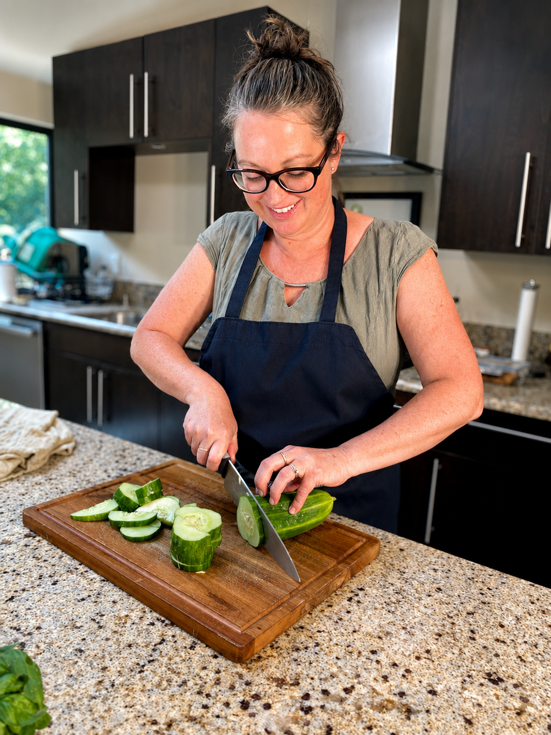 A woman with glasses and a bun, wearing a grey shirt and a navy apron, is slicing a cucumber on a wooden cutting board in a kitchen.