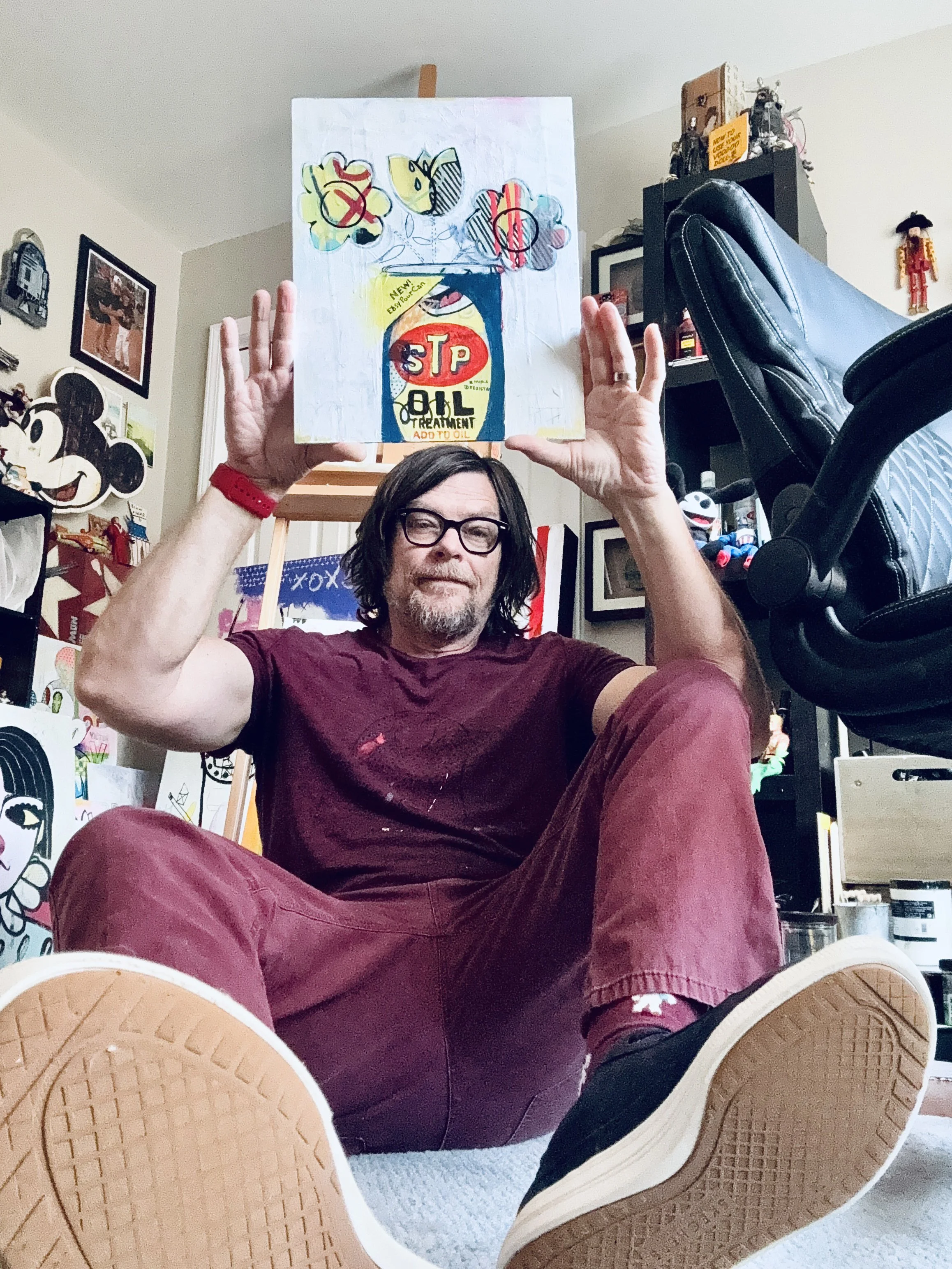 A man with glasses, a beard, and long hair is sitting on the floor, holding a colorful artwork above his head. He is wearing a maroon shirt, maroon pants, and sneakers. The room around him is decorated with various art and memorabilia, including Mickey Mouse and other items on the walls and shelves.