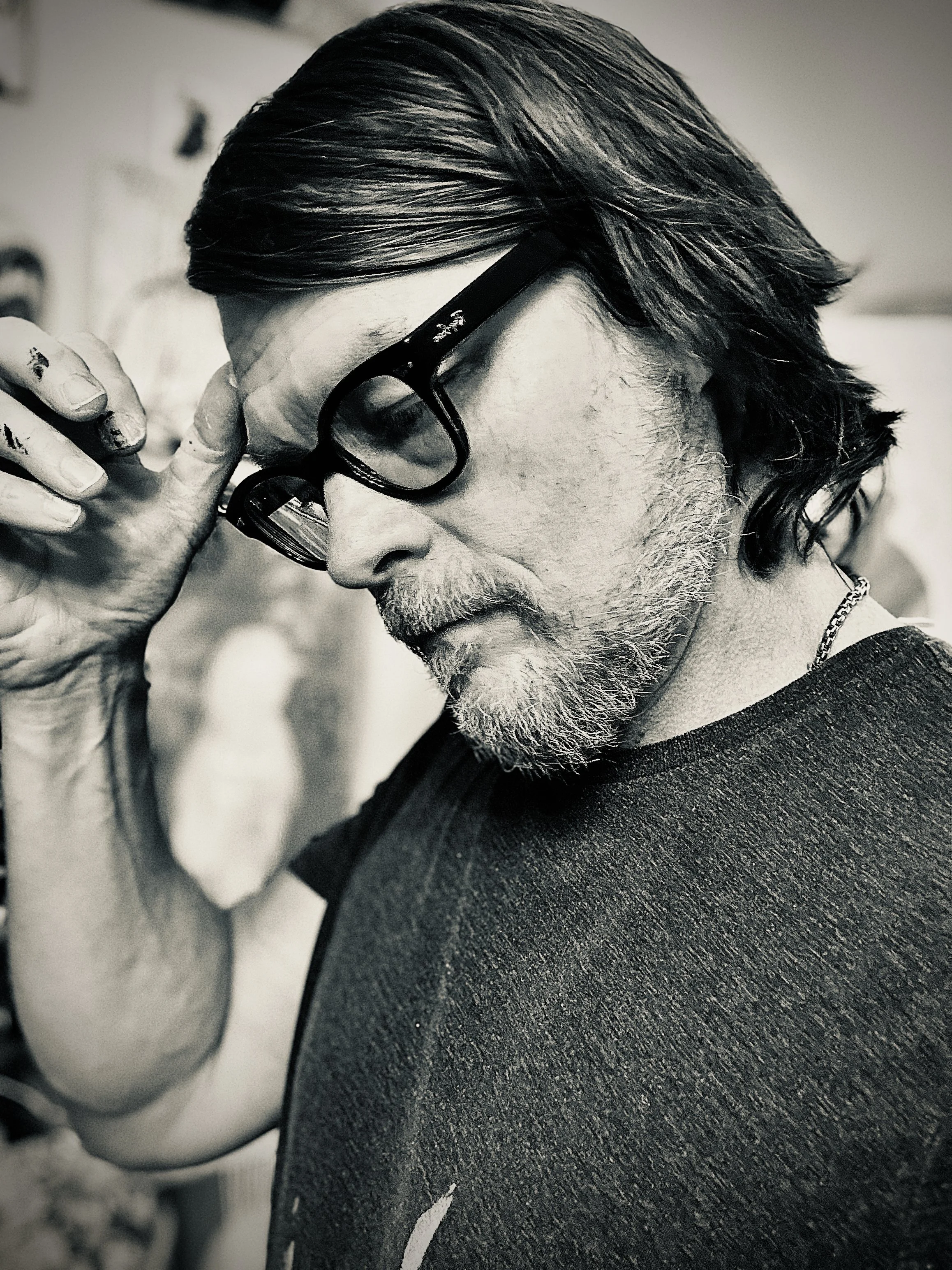 Close-up of a man with dark, styled hair and glasses, resting his finger on his lips, wearing a dark shirt and a chain necklace.