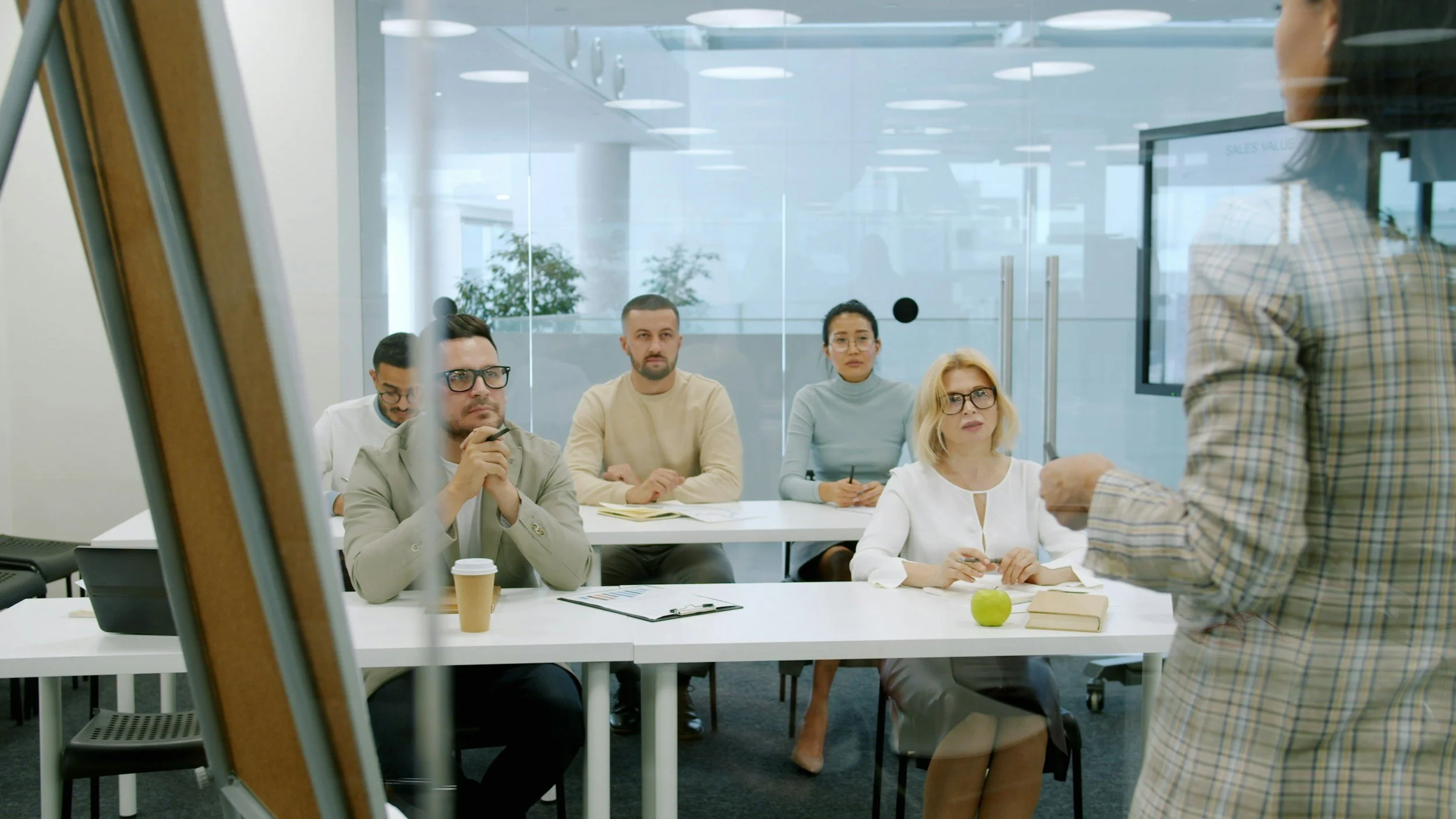 A group of five diverse adults attending a business meeting or workshop in a modern conference room, listening to a speaker standing at the front.