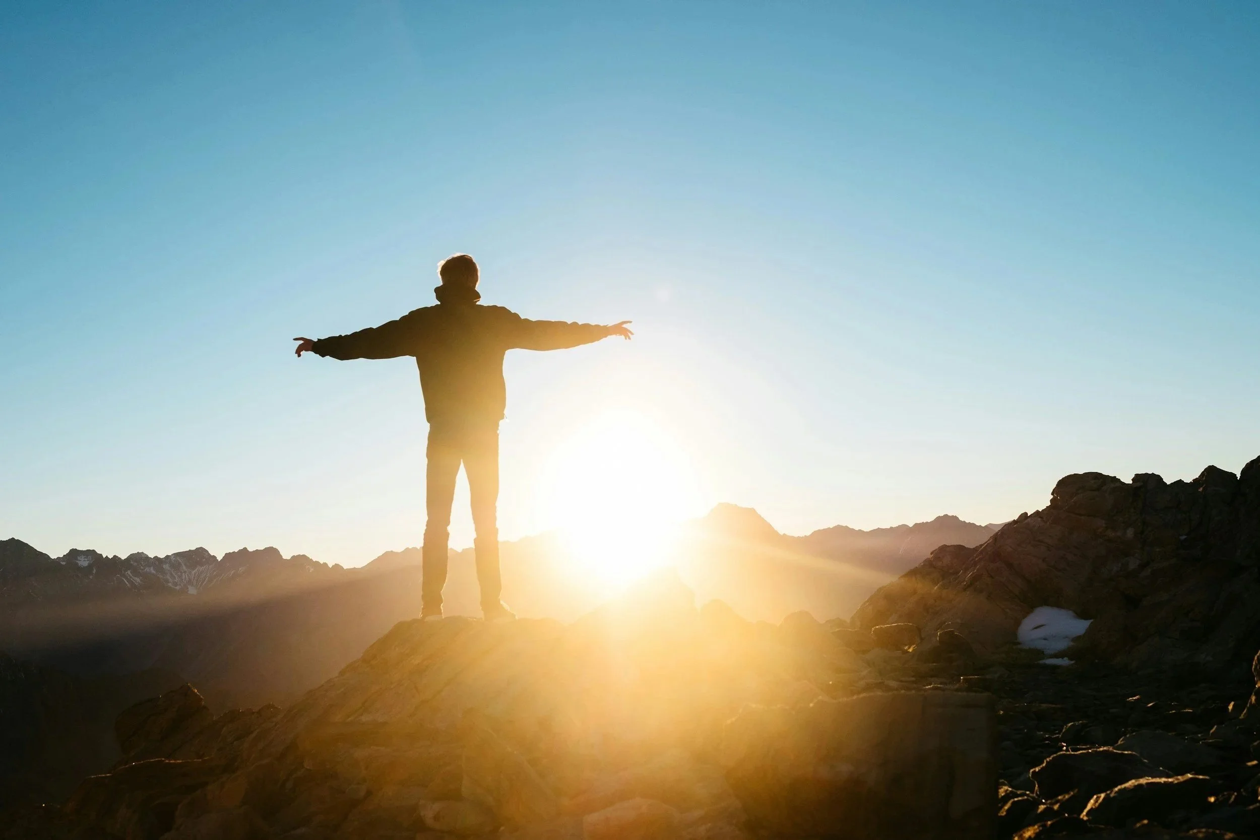 Person standing on mountain ridge with arms outstretched, facing the sunrise or sunset over a mountain landscape.
