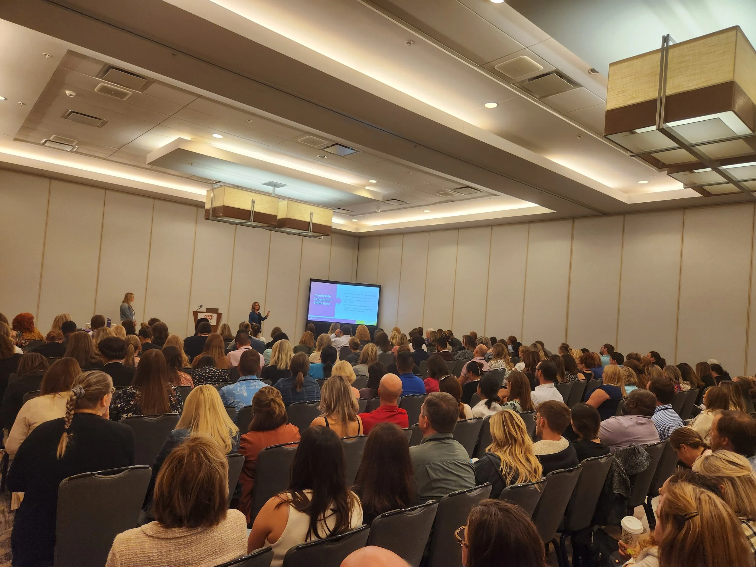 A large conference room filled with seated attendees watching a woman presenting on stage with a screen displaying a presentation slide.