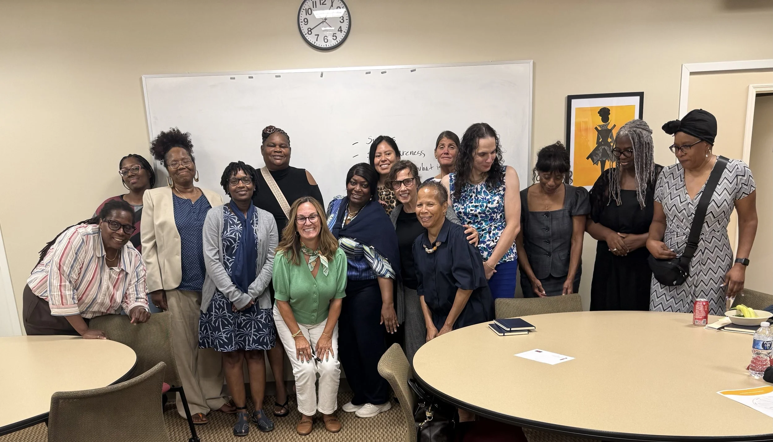 A group of diverse women standing together in a conference room, smiling at the camera. There are round tables with chairs, a whiteboard with written notes, and framed art on the walls. A clock shows the time as 8:11.