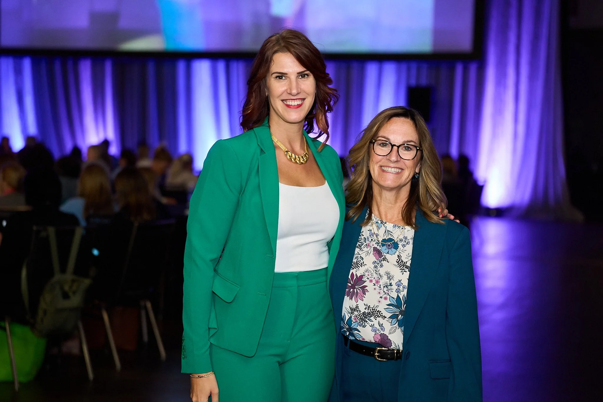 Two women, one taller in a green suit with auburn hair and one shorter with glasses in a turquoise suit with flowered blouse and longer brown hair