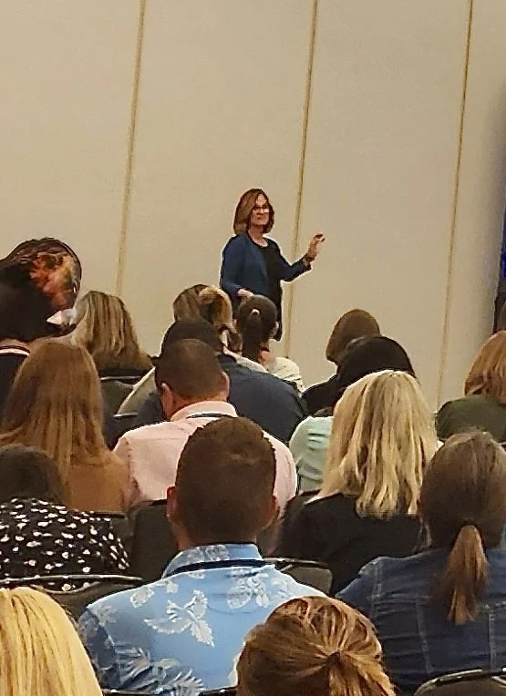 A woman standing in front of a room of seated audience members, giving a presentation or speech.