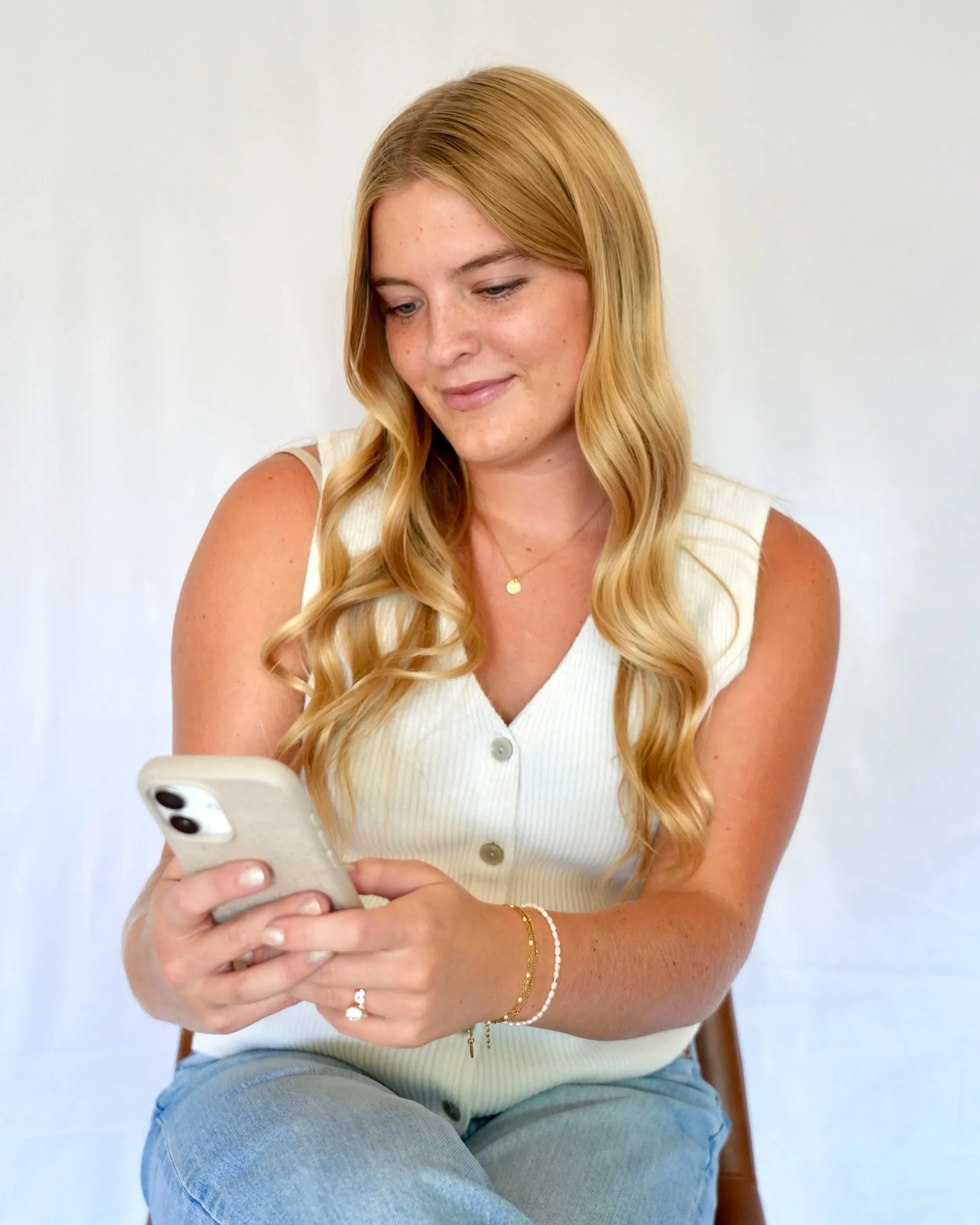 A young woman with long, wavy blonde hair, wearing a sleeveless white top and blue jeans, sitting on a wooden chair, looking at her smartphone with a slight smile.
