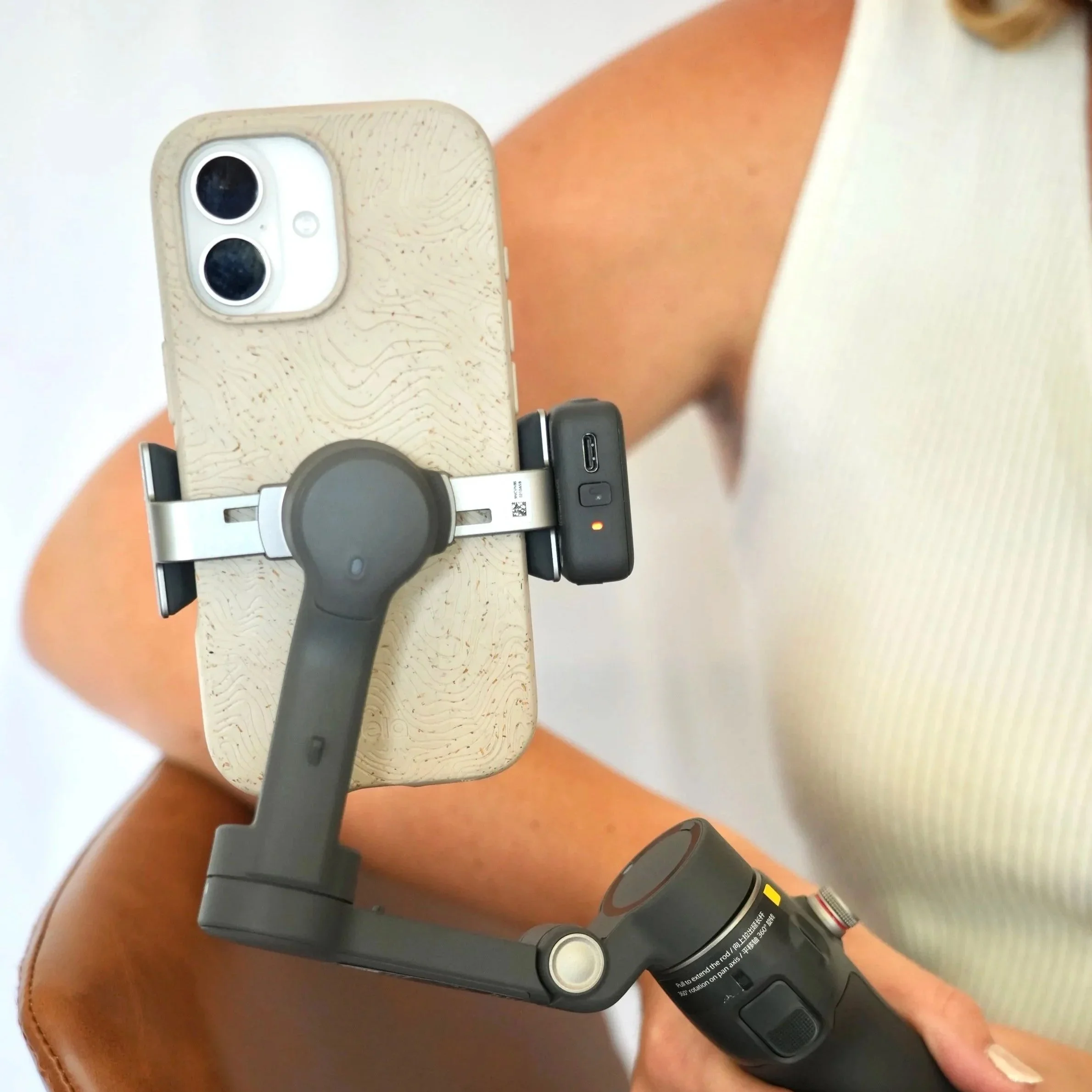 A person's hand holding a wireless remote control connected to a device, with a smartphone mounted on it, on a light brown surface.