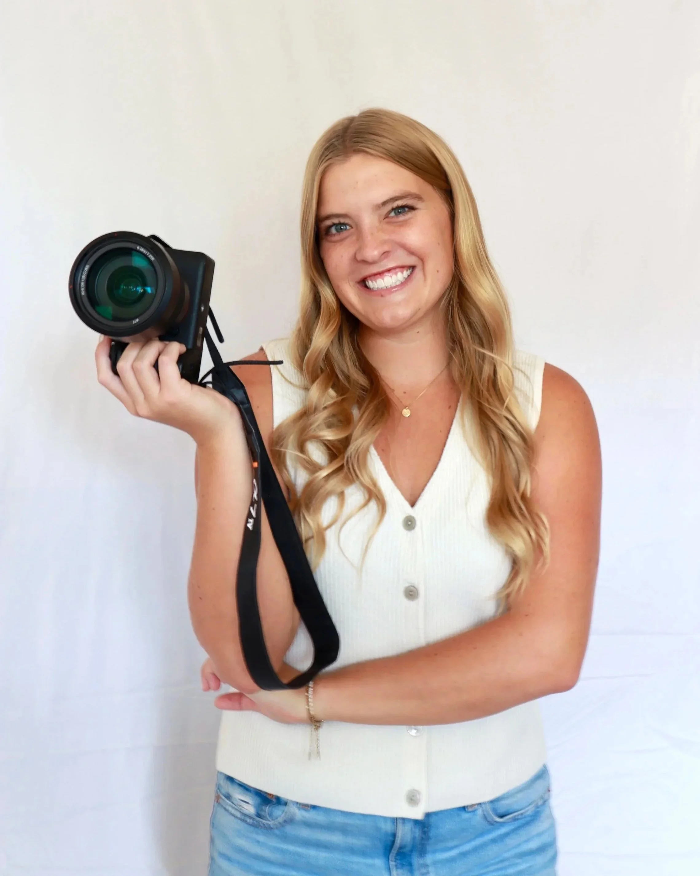 A young woman with long blonde hair, smiling, holding a camera with her left hand, standing in front of a plain white wall, wearing a white sleeveless button-up top and jeans.