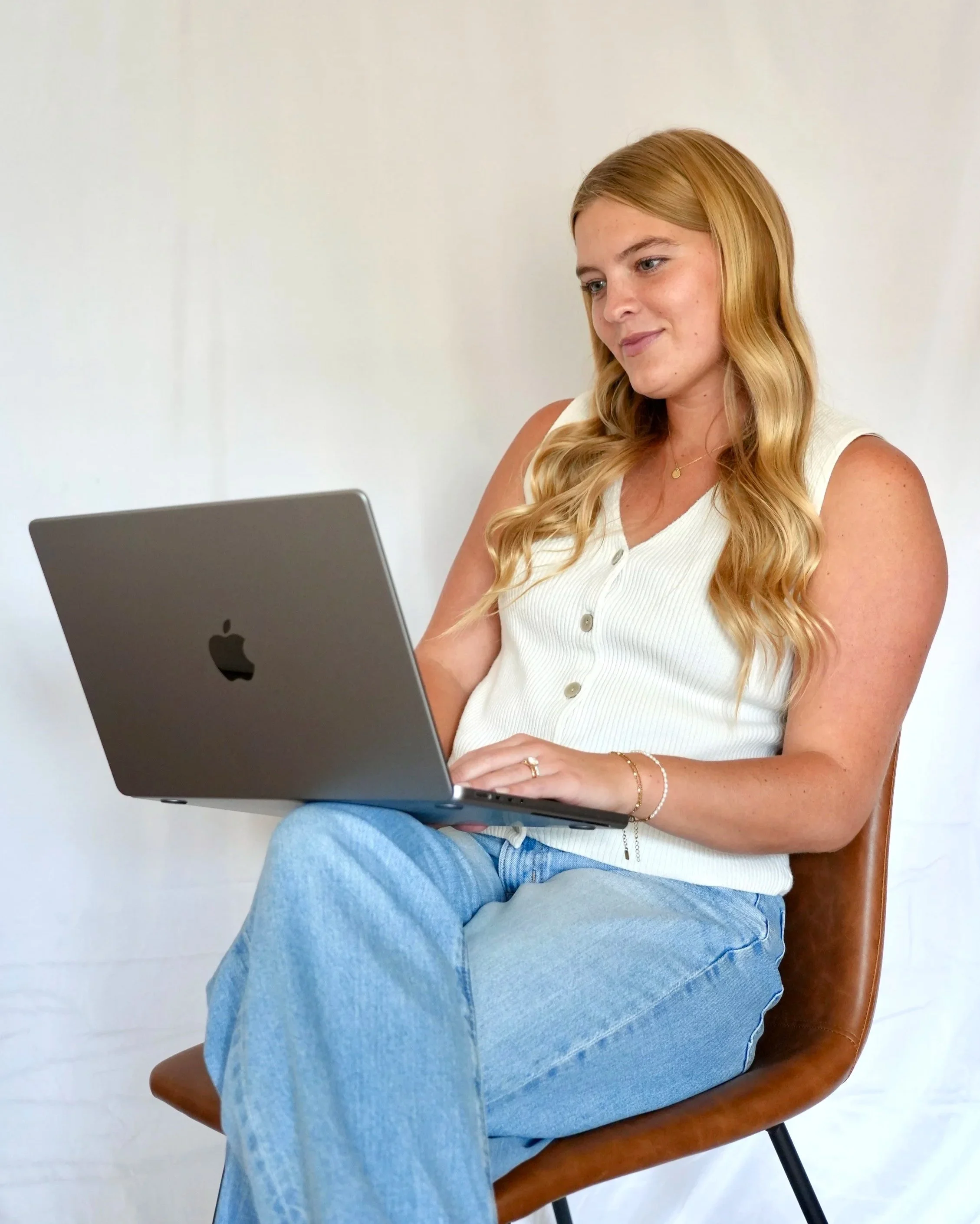 A woman with long blonde hair sitting on a wooden chair, looking at a laptop, against a plain white wall background.