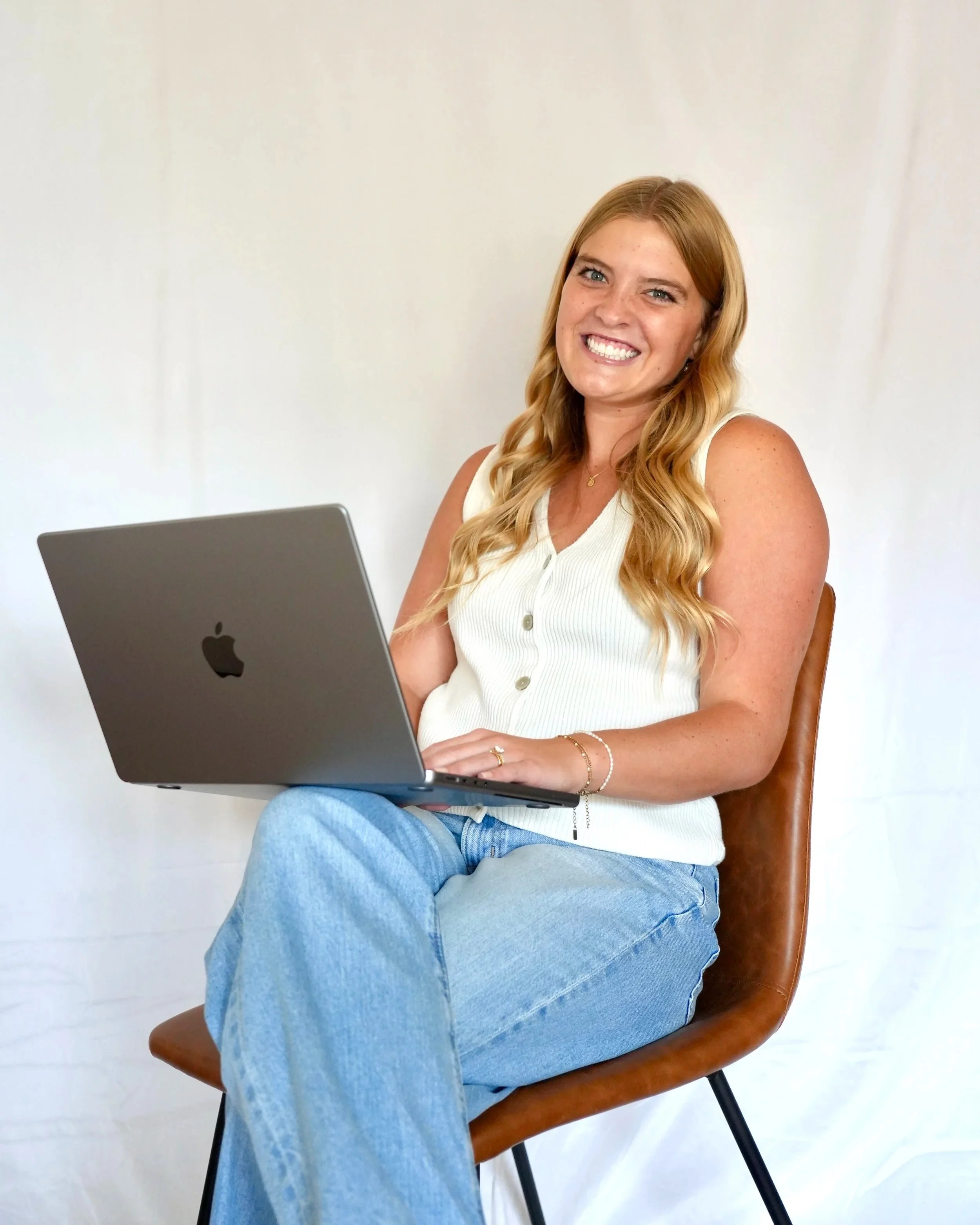 A woman with long blonde hair, wearing a white sleeveless top and light blue jeans, sits on a brown chair in front of a plain white background, smiling at the camera with a silver MacBook on her lap.