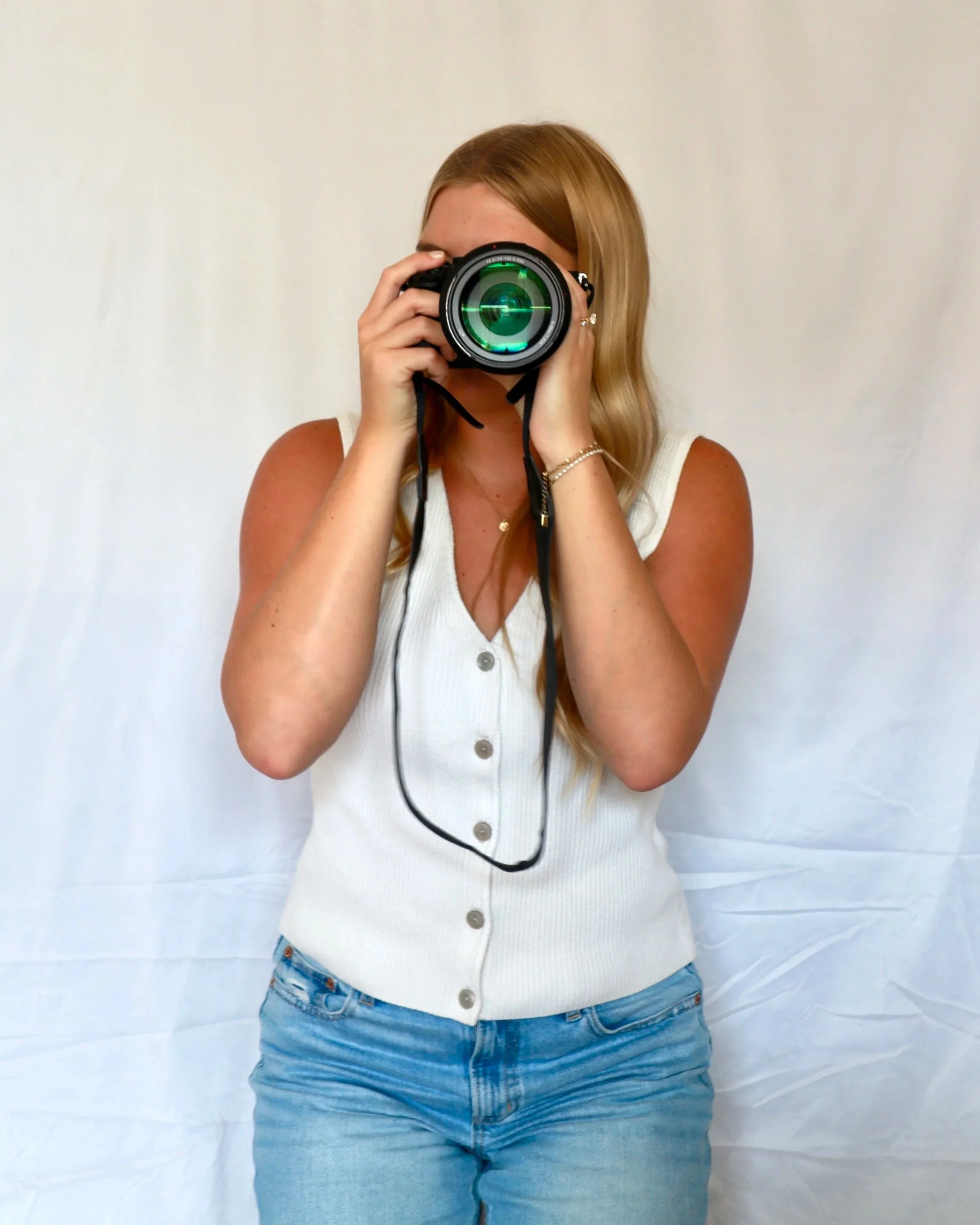Woman with long blonde hair wearing a white sleeveless top with buttons and blue jeans, holding a camera up to her face, standing in front of a plain white background.