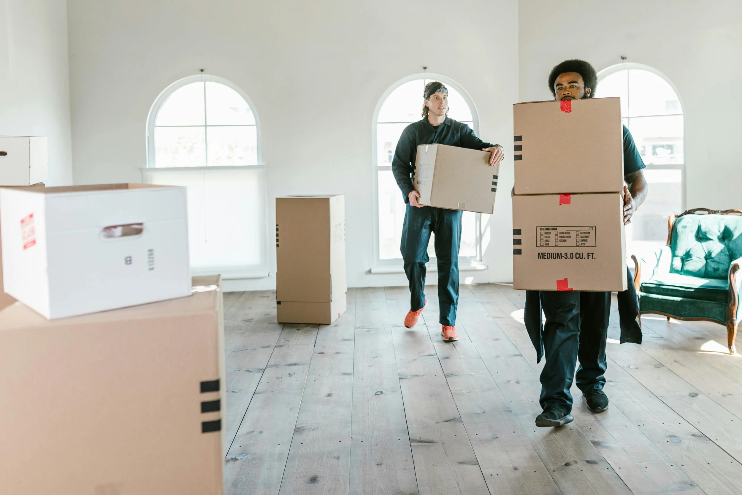 Two men are moving boxes inside a brightly lit room with arched windows. One man with an afro hairstyle and a beard is carrying a large box, while the other man with long hair and a headband carries a smaller box. Several cardboard boxes are scattered around.