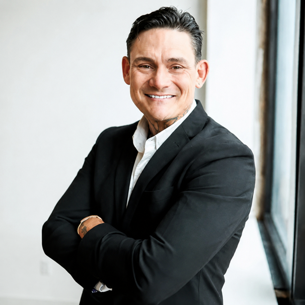 A smiling man in a black suit with white shirt, arms crossed, standing near a window.