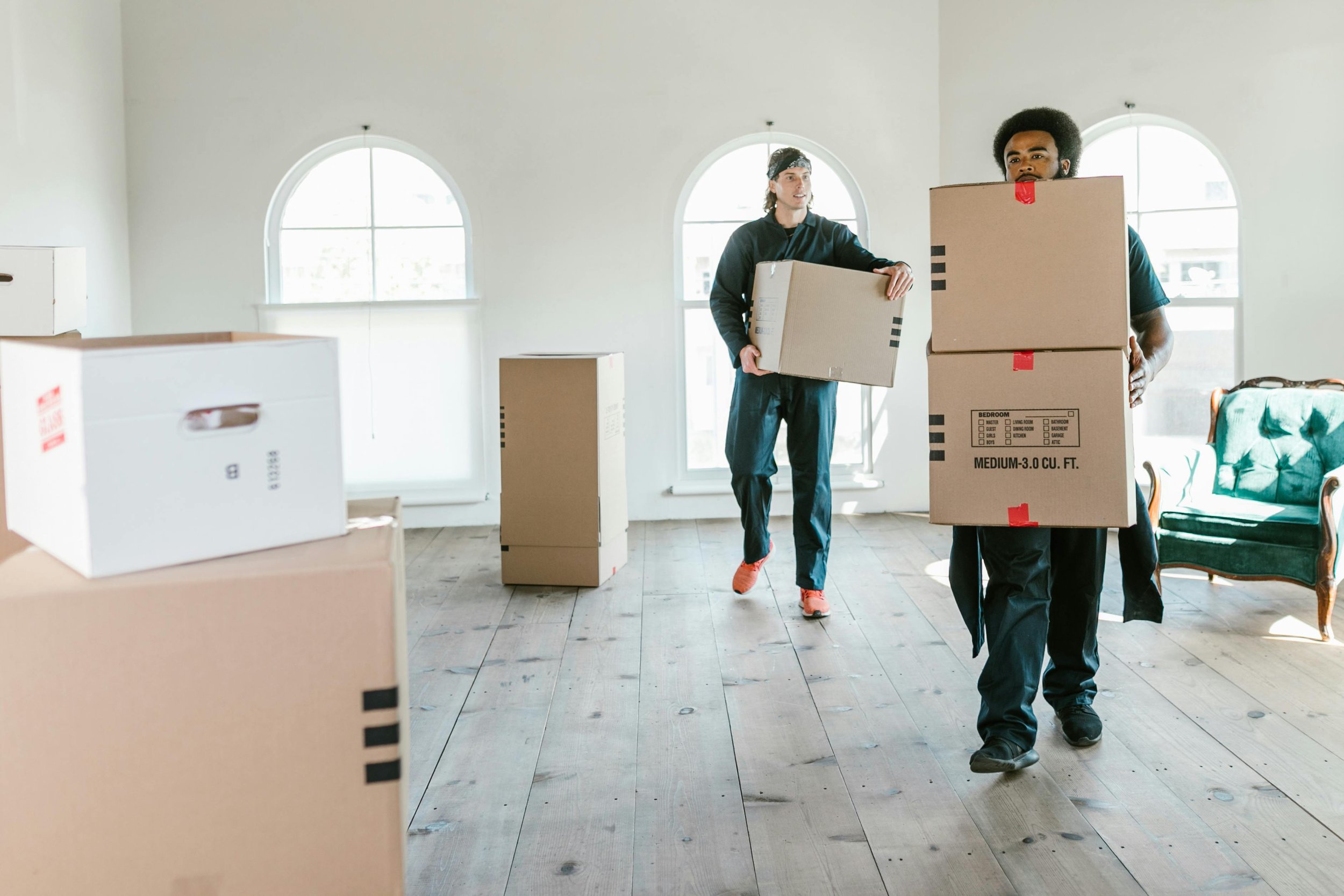 Two men carrying moving boxes inside a bright room with large arched windows. One man has curly hair and a beard, wearing a dark shirt, while the other man has shoulder-length hair and a headband. There are additional boxes and a green chair with wooden arms visible in the room.