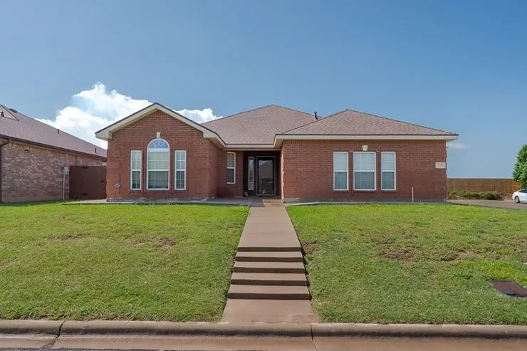 Single-story brick house with a front porch, located in a grassy yard, under a clear blue sky.
