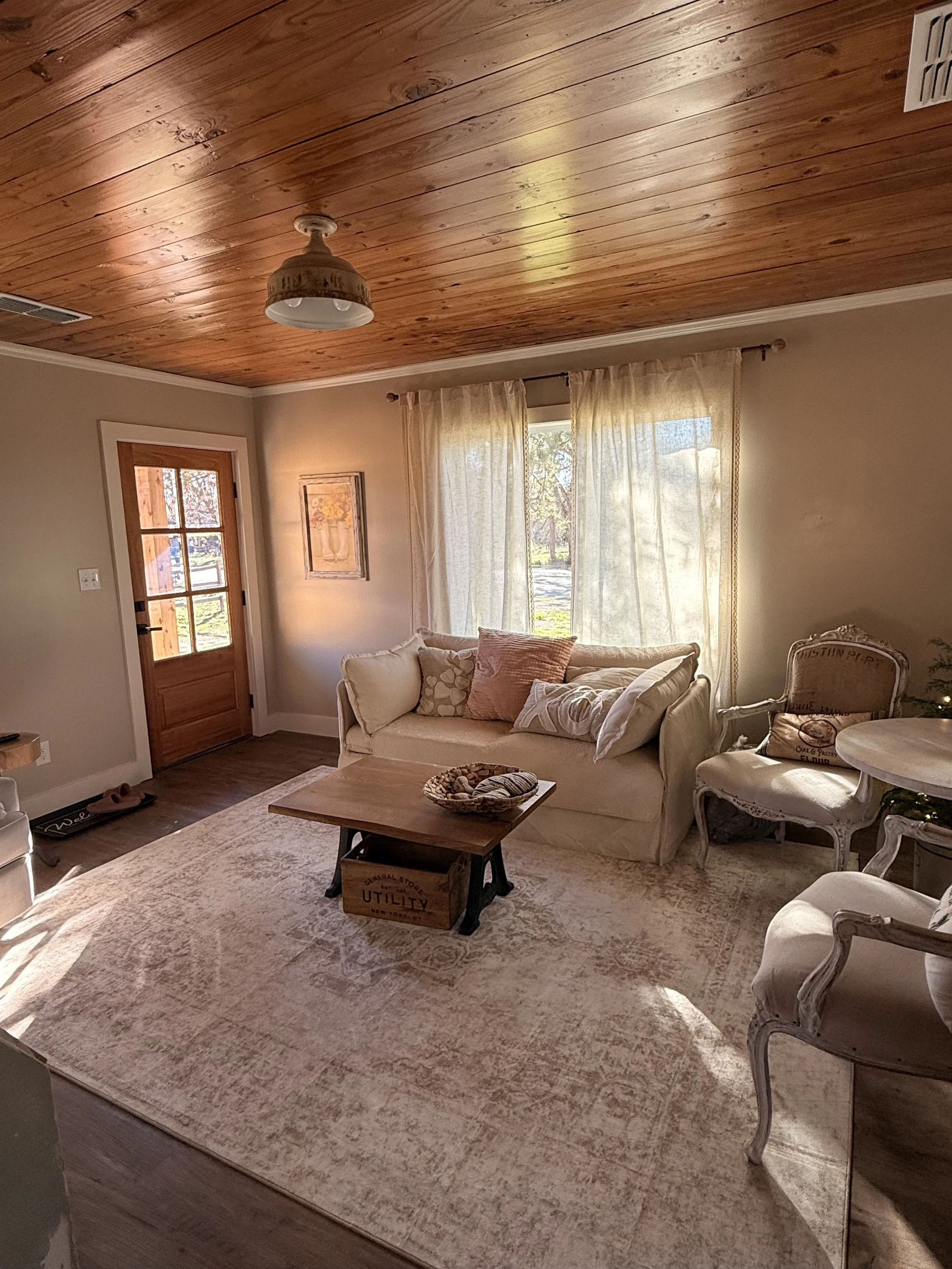 A cozy living room with a wooden ceiling, beige walls, and large window with sheer curtains. There is a white sofa with throw pillows, a vintage armchair, a wooden coffee table with a basket, and a rug.