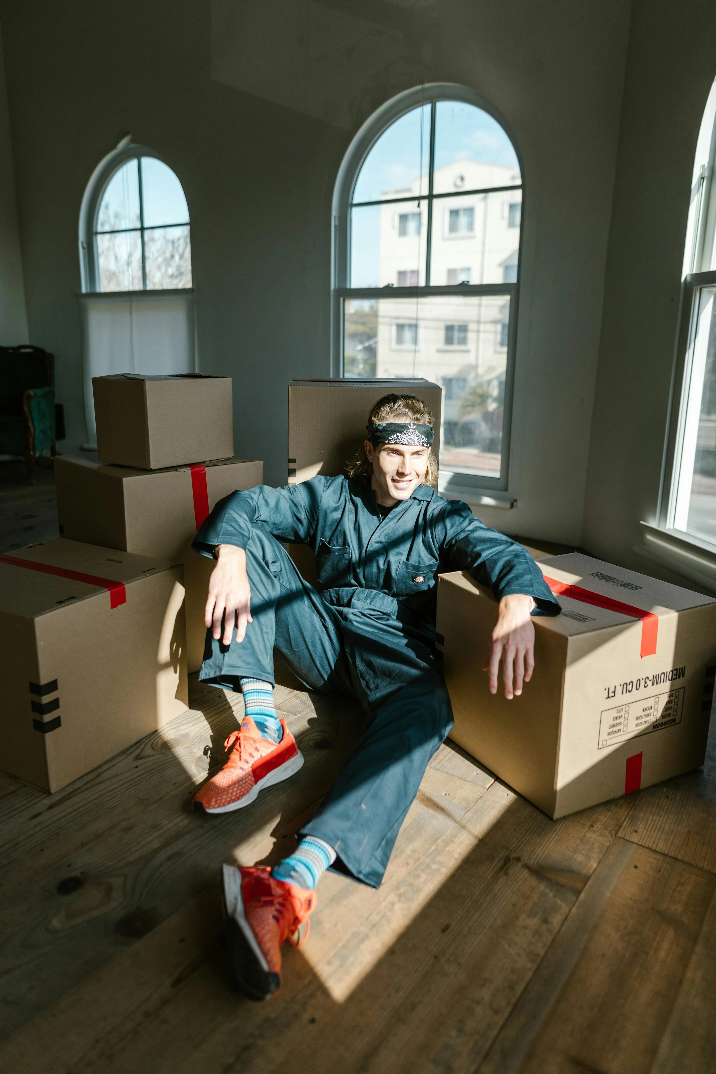 A young man in a blue jumpsuit and colorful sneakers sits on the wooden floor of a sunlit room, surrounded by cardboard boxes, smiling at the camera with large arched windows behind him.