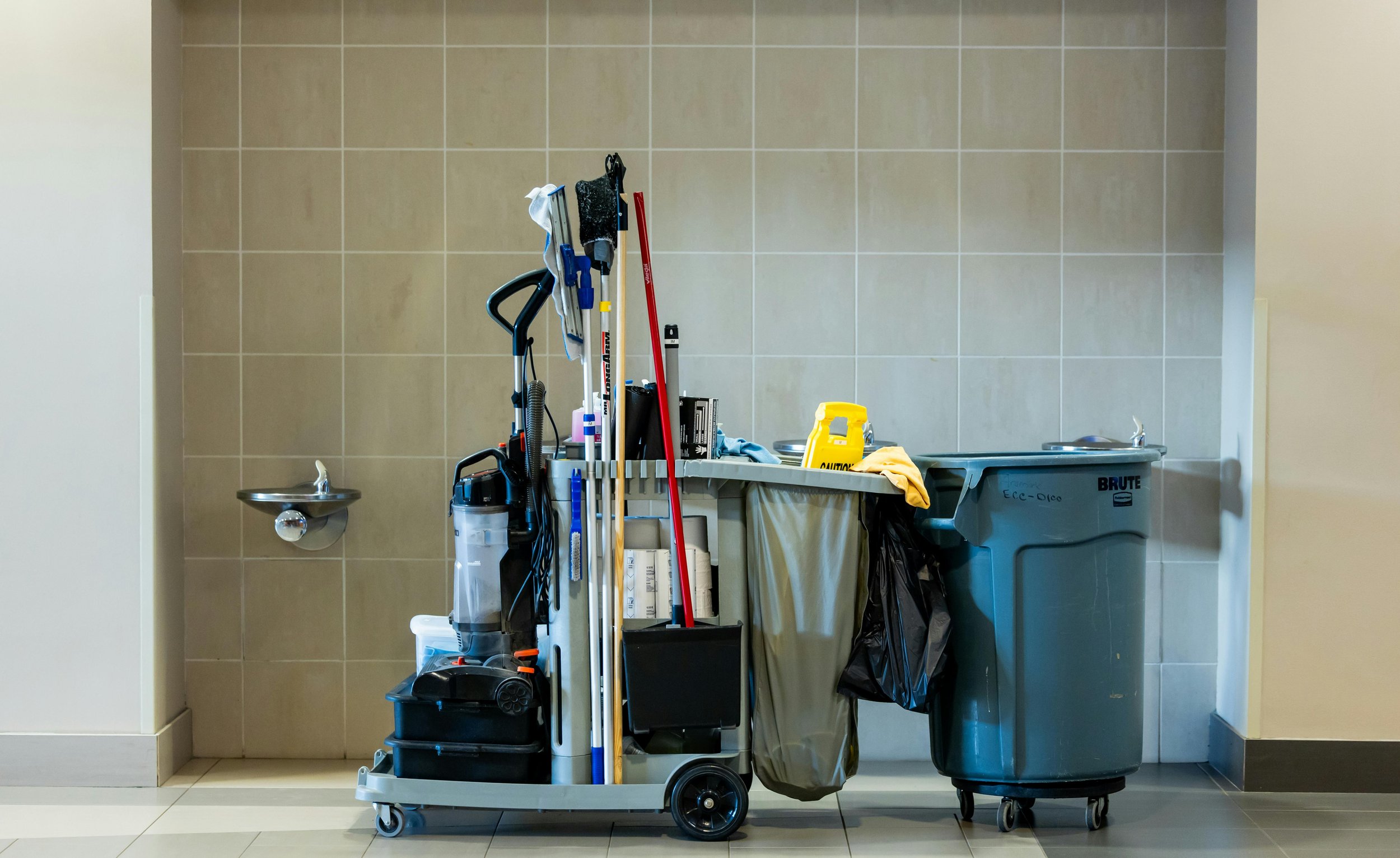 Cleaning supplies and equipment stacked on a cart, with a trash can and a sink in the background, against a beige tiled wall.