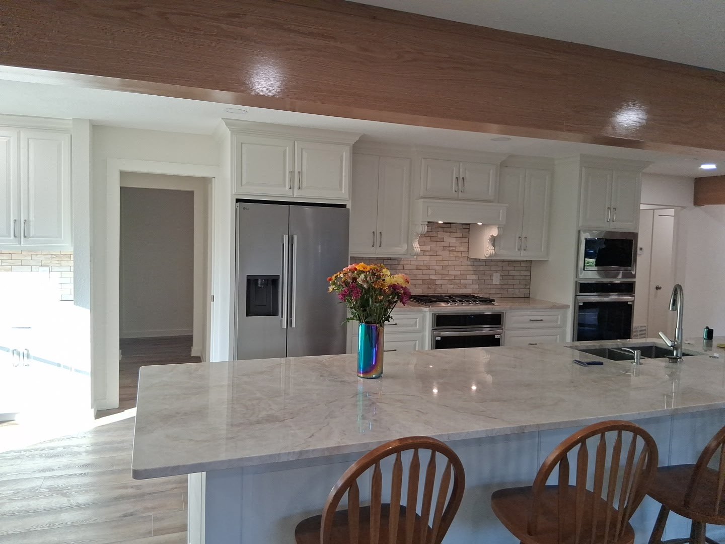 Modern kitchen with white cabinets, stainless steel appliances, and a marble island with a vase of colorful flowers, wooden chairs, and a brick backsplash.