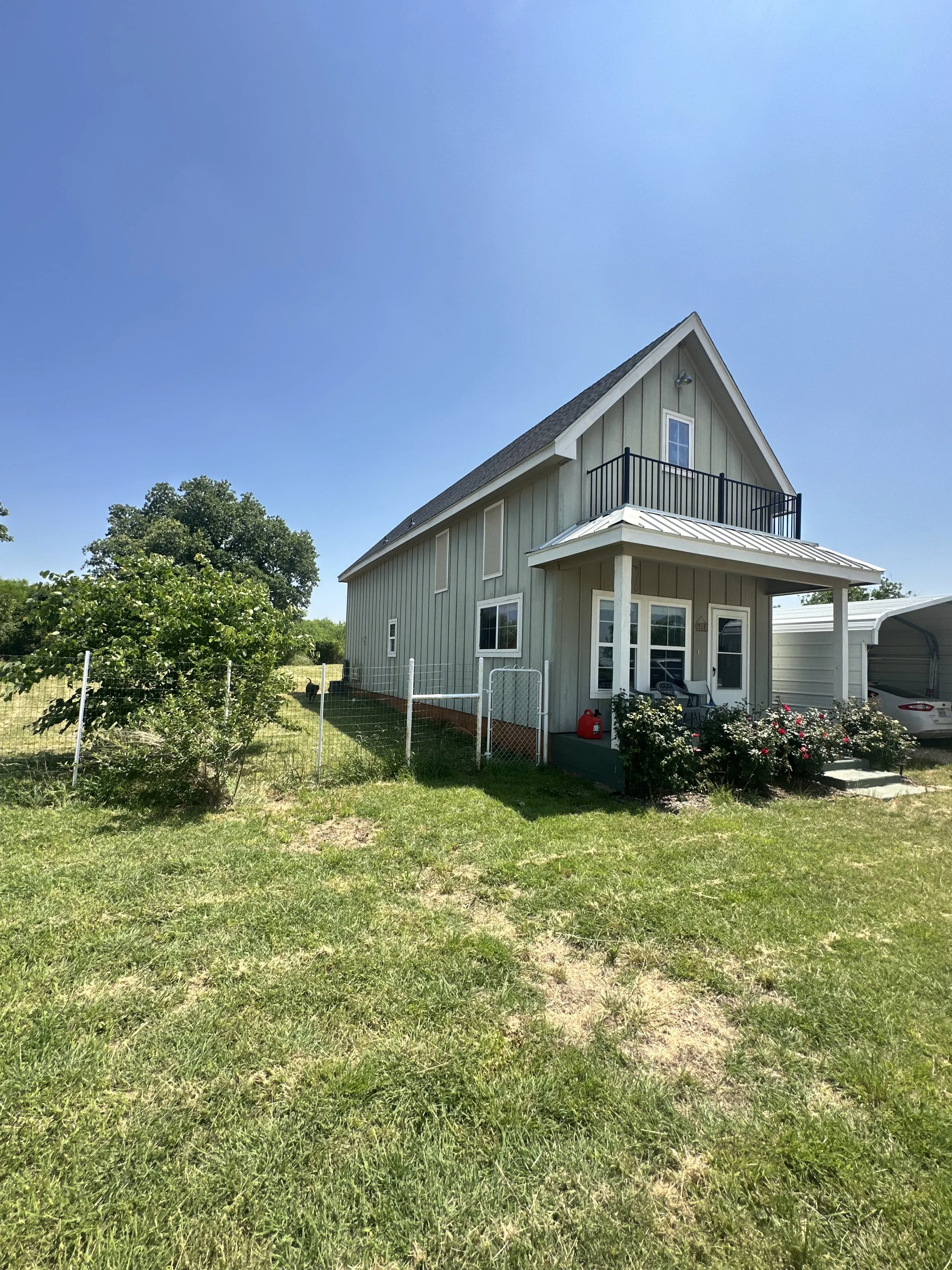 A two-story house with gray siding and a metal roof, surrounded by a grassy yard with a small fence, green trees, and a clear blue sky.