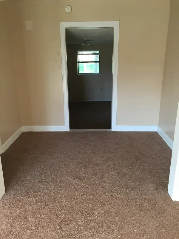 View of an empty room with beige walls, brown carpet, and an open door leading to another room with a window.