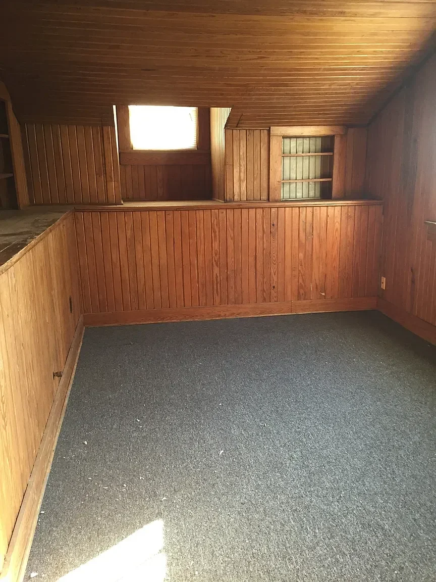 A small room with wood-paneled walls and ceiling, a window on the back wall, and a dark carpeted floor with some debris.