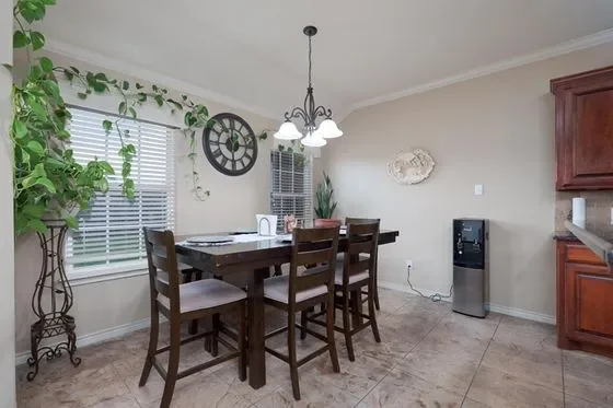 Dining room with a wooden table, six chairs, a chandelier, a wall clock, window blinds, a water dispenser, and kitchen cabinets.
