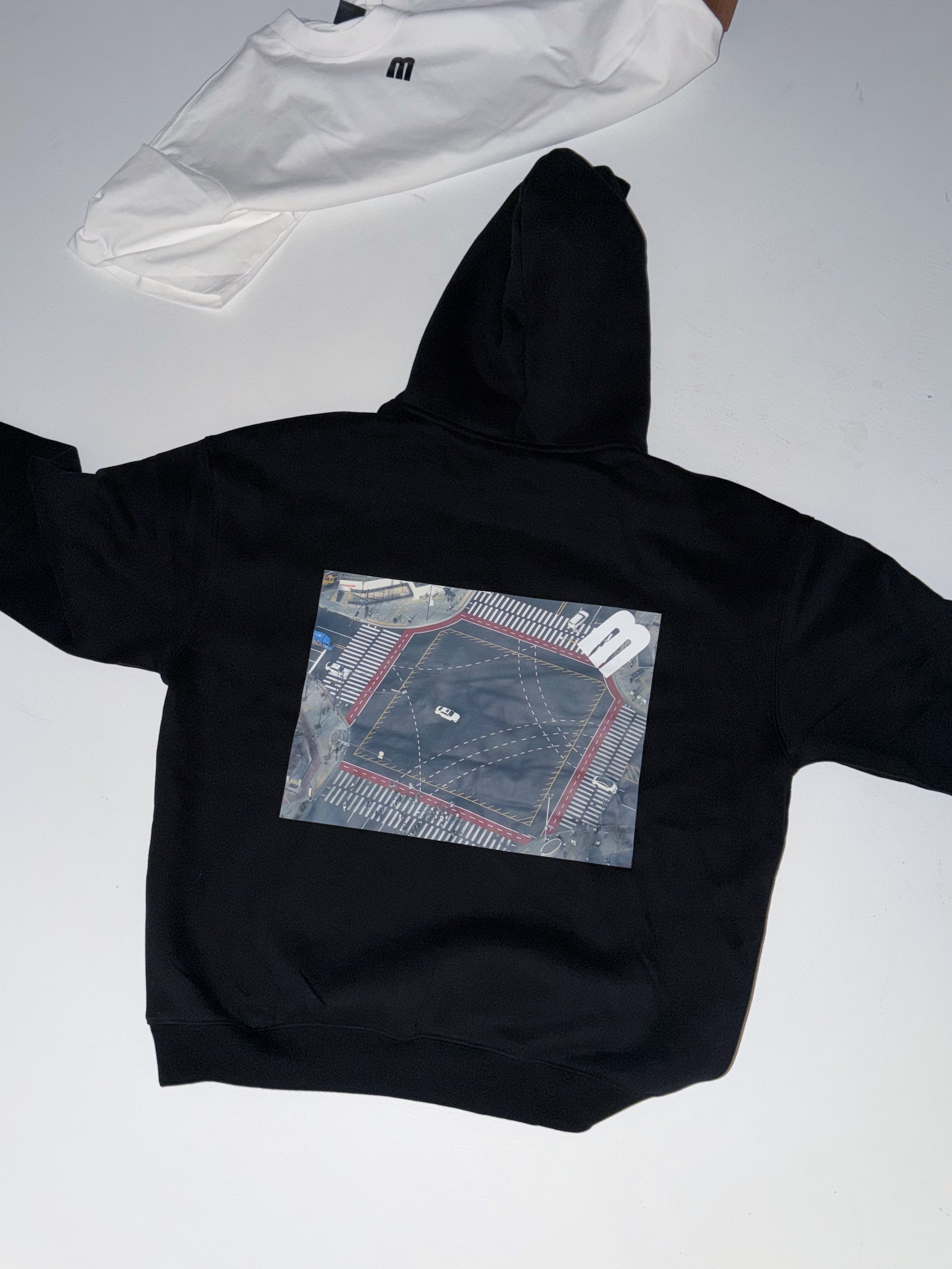A person wearing a black hoodie with a graphic print on the front, standing against a white background, with white and black clothing items or accessories nearby.