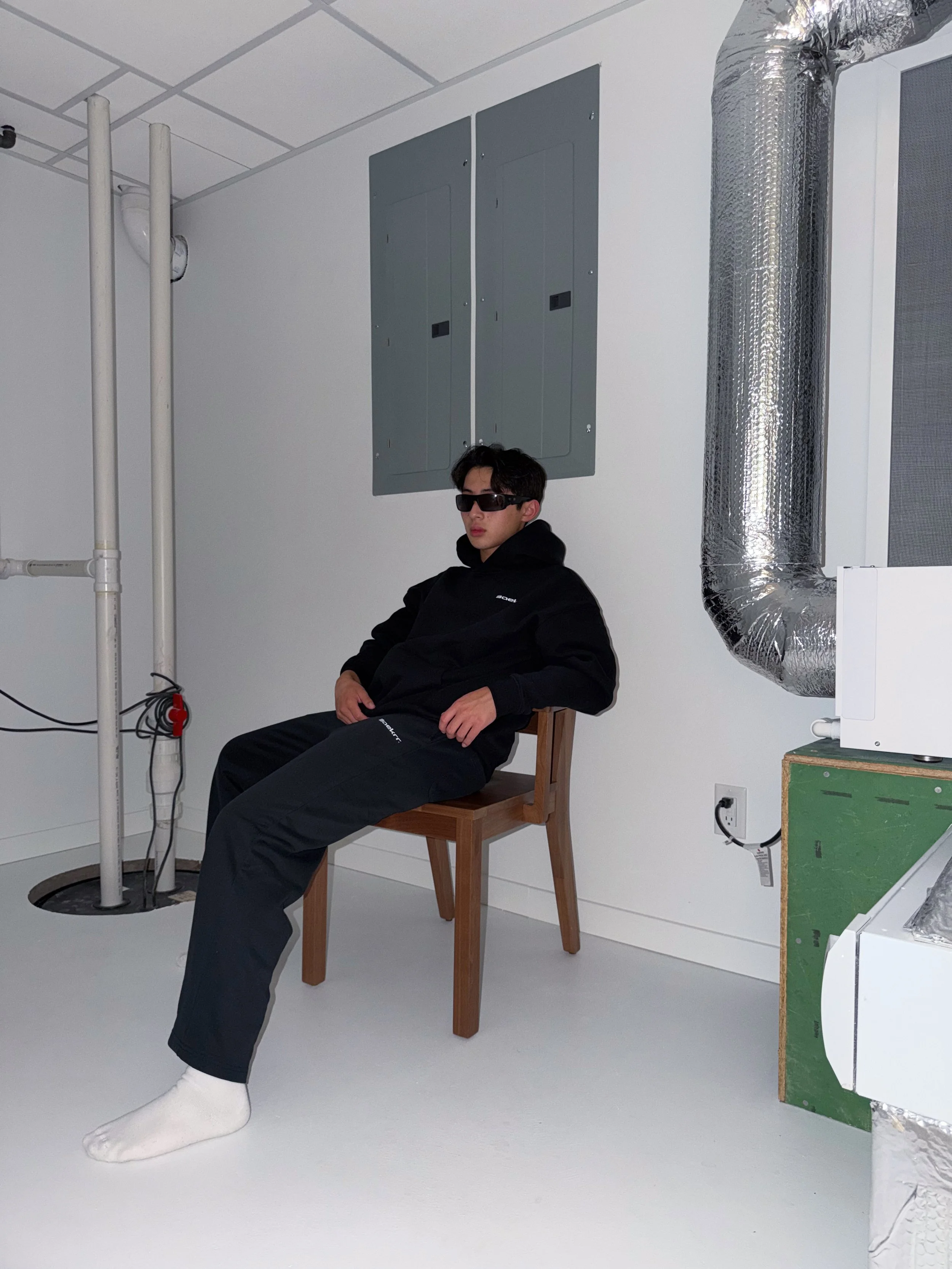 A young man wearing black sunglasses, a black hoodie, black pants, and white socks sitting on a wooden chair in a room with white walls and ceiling tiles, next to a large silver ventilation pipe, electrical panels, and an area with wires and a power 