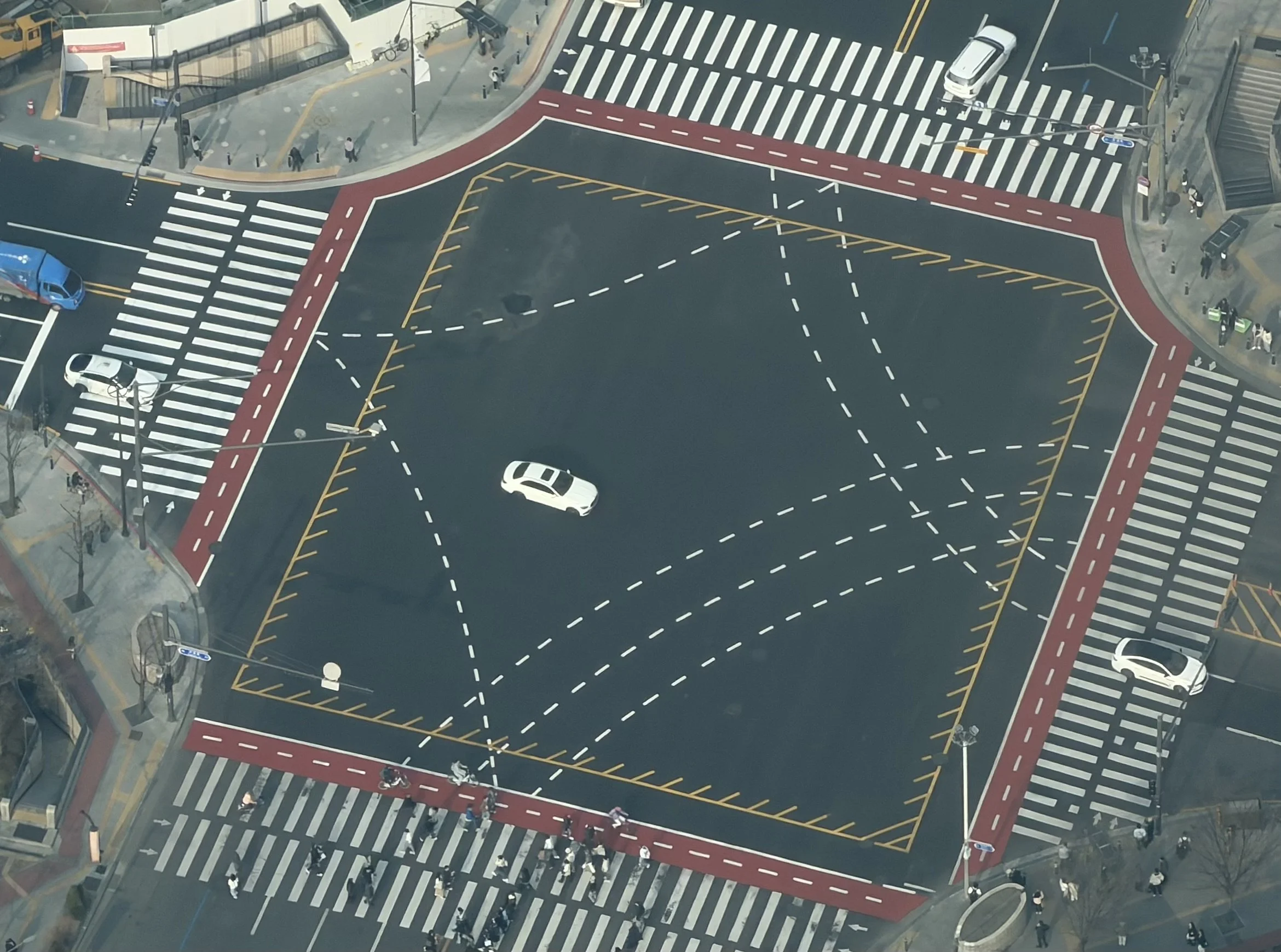 An aerial view of a busy city intersection with multiple crosswalks, cars waiting at traffic signals, and pedestrians crossing the streets. The intersection includes designated bike lanes and a bus stop nearby.