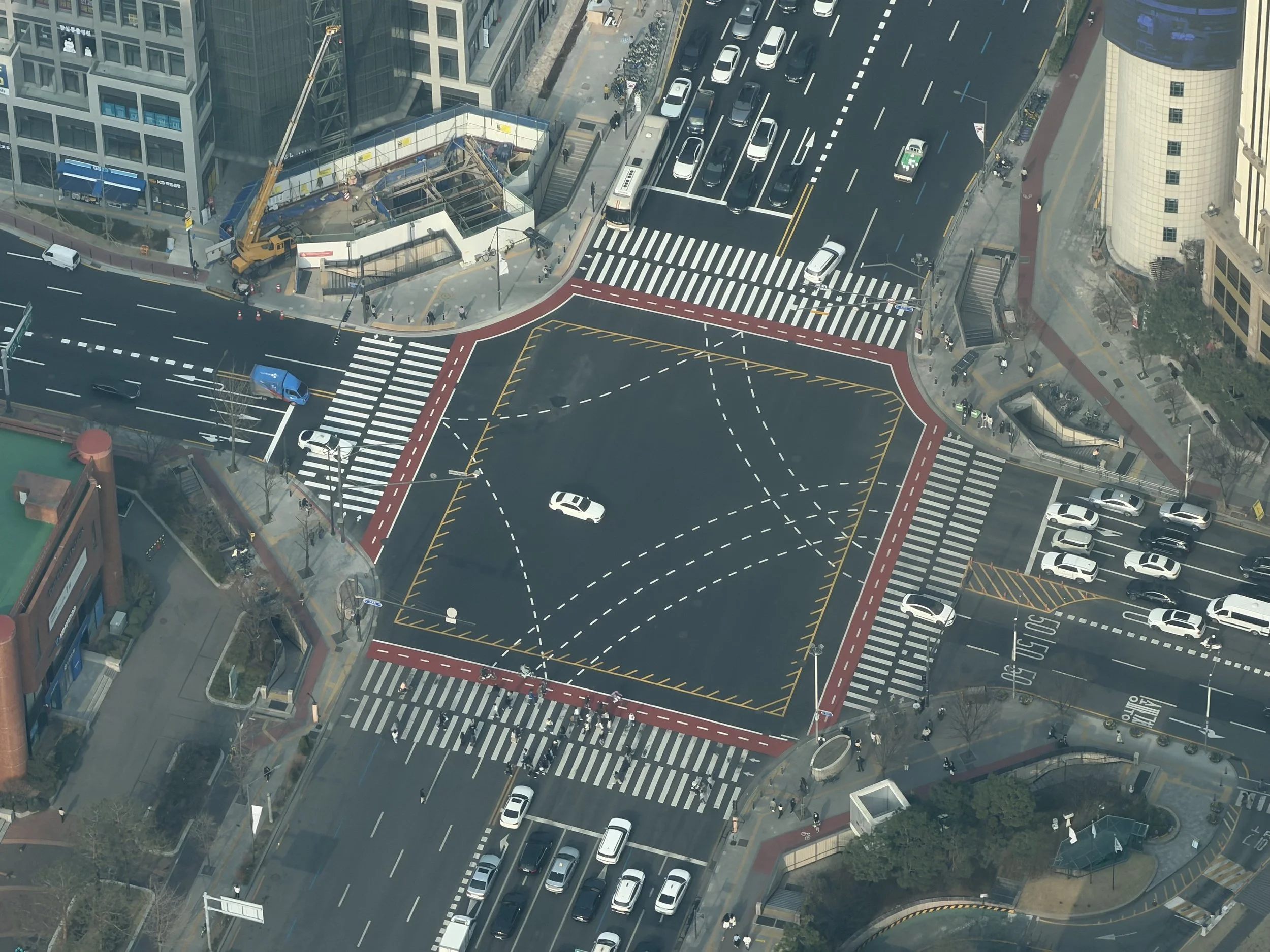 An aerial view of a busy city street intersection with marked crosswalks, cars, buses, a construction site with a crane, and surrounding tall buildings.