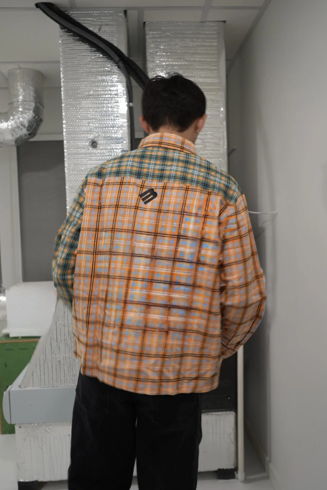 Back view of a person in a colorful plaid shirt standing in an industrial or refrigerated kitchen area, possibly near a freezer or walk-in cooler.
