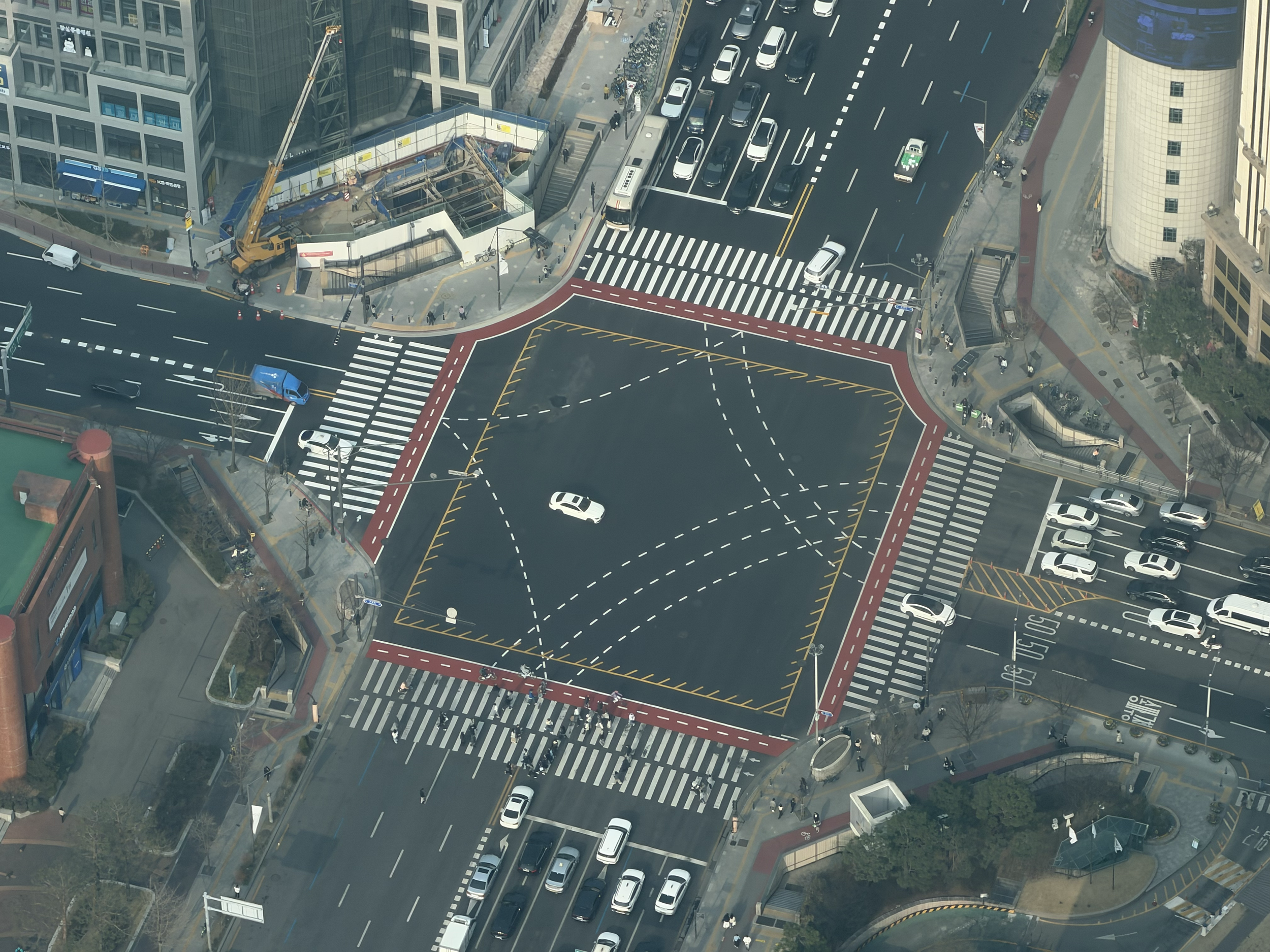 An aerial view of a city street intersection with crosswalks, traffic lanes, vehicles including cars and buses, and construction work on a building site.