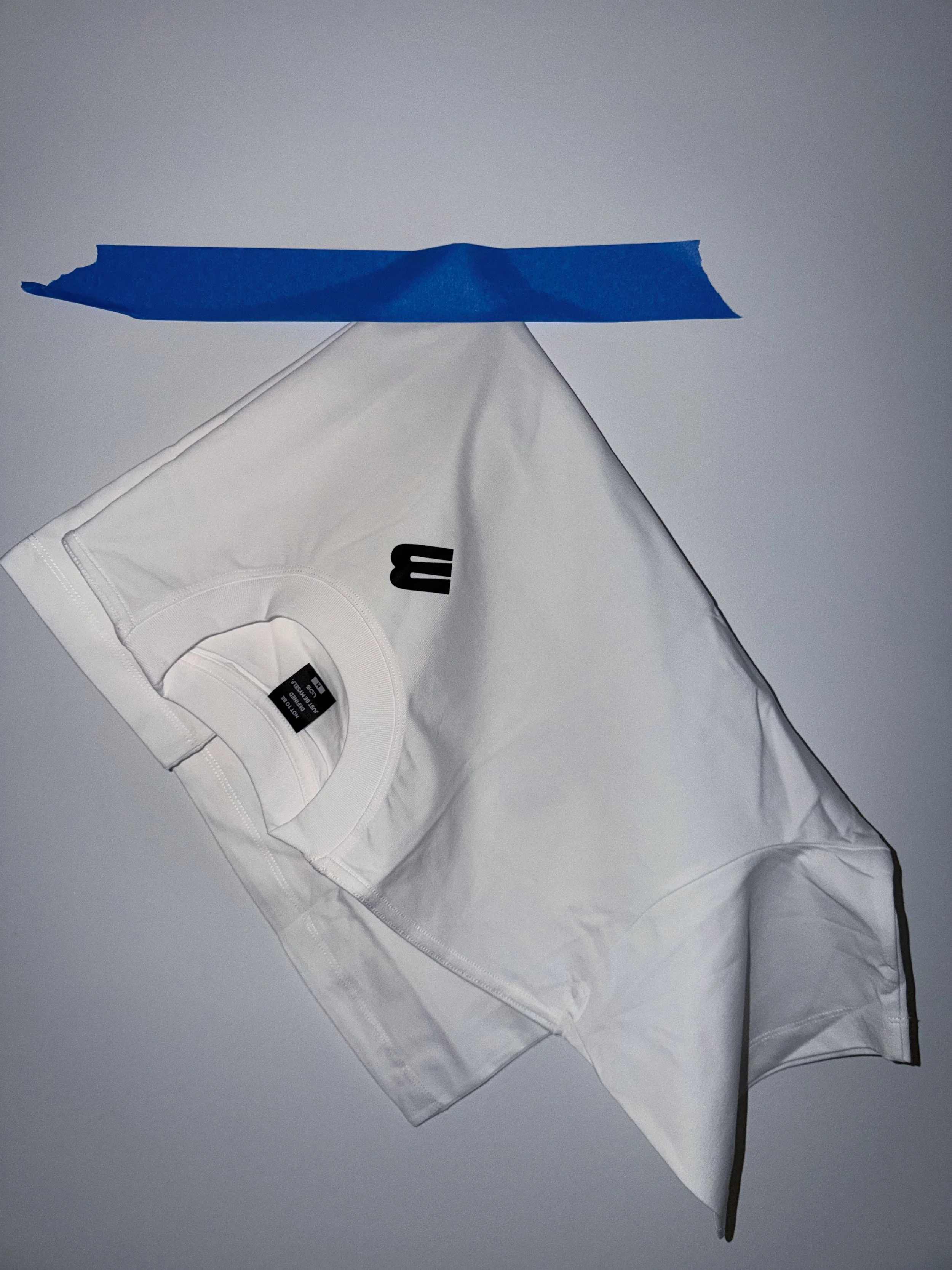 A white piece of clothing, possibly a shirt, on a flat surface, with a black tag visible inside and a black cord attached, along with a small piece of blue painter's tape placed on top.