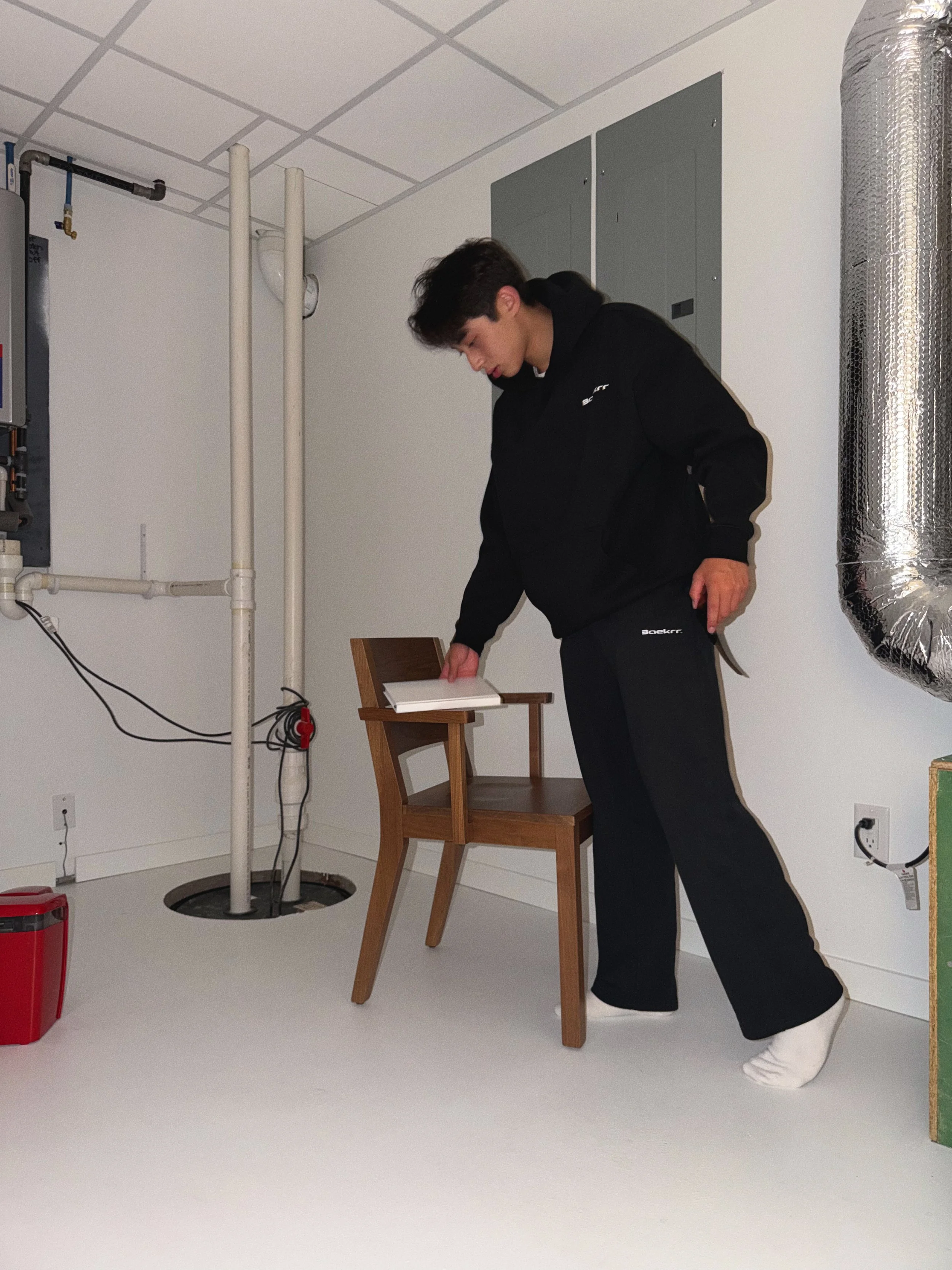 A man leaning over a small wooden table with a white book on top, inside a room with white walls, a green electrical panel, and a silver duct on the right. There is a chair in front of the table and various electrical components and pipes on the wall